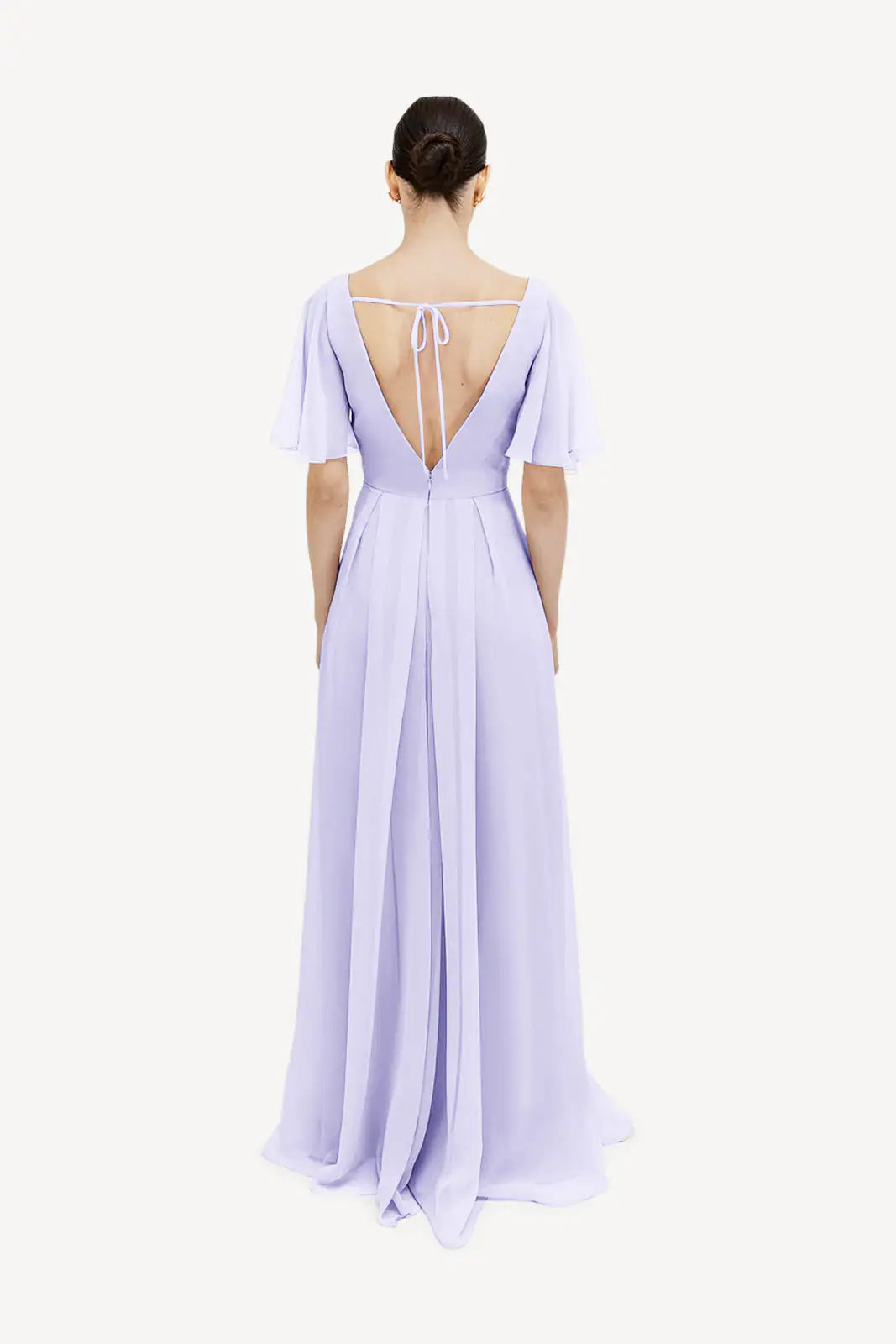 A woman with dark hair in a low bun wears the TH&TH Phoebe Bridesmaid Dress in Lilac Mist chiffon, featuring flutter sleeves and a deep V-back with thin straps, elegantly showcasing the gown’s back details as she stands facing away.