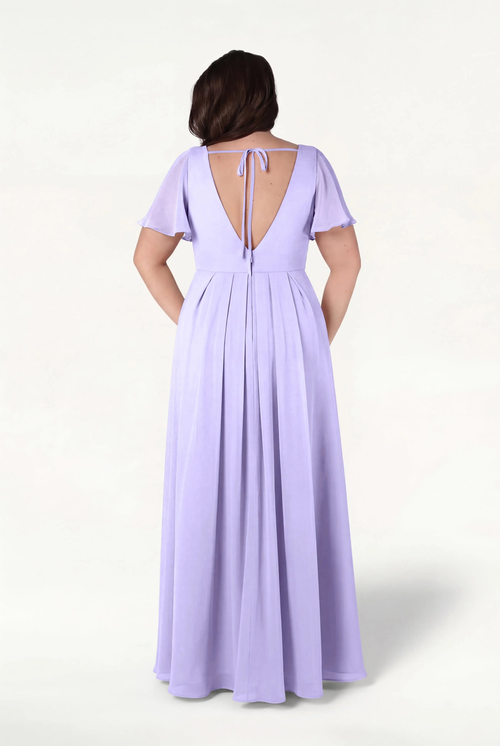 A woman stands with her back to the camera in a TH&TH Phoebe Bridesmaid Dress in Lilac Mist Chiffon, featuring a floor-length silhouette, short flutter sleeves, and a deep V-back tied with a bow—an effortlessly elegant bump-friendly choice.