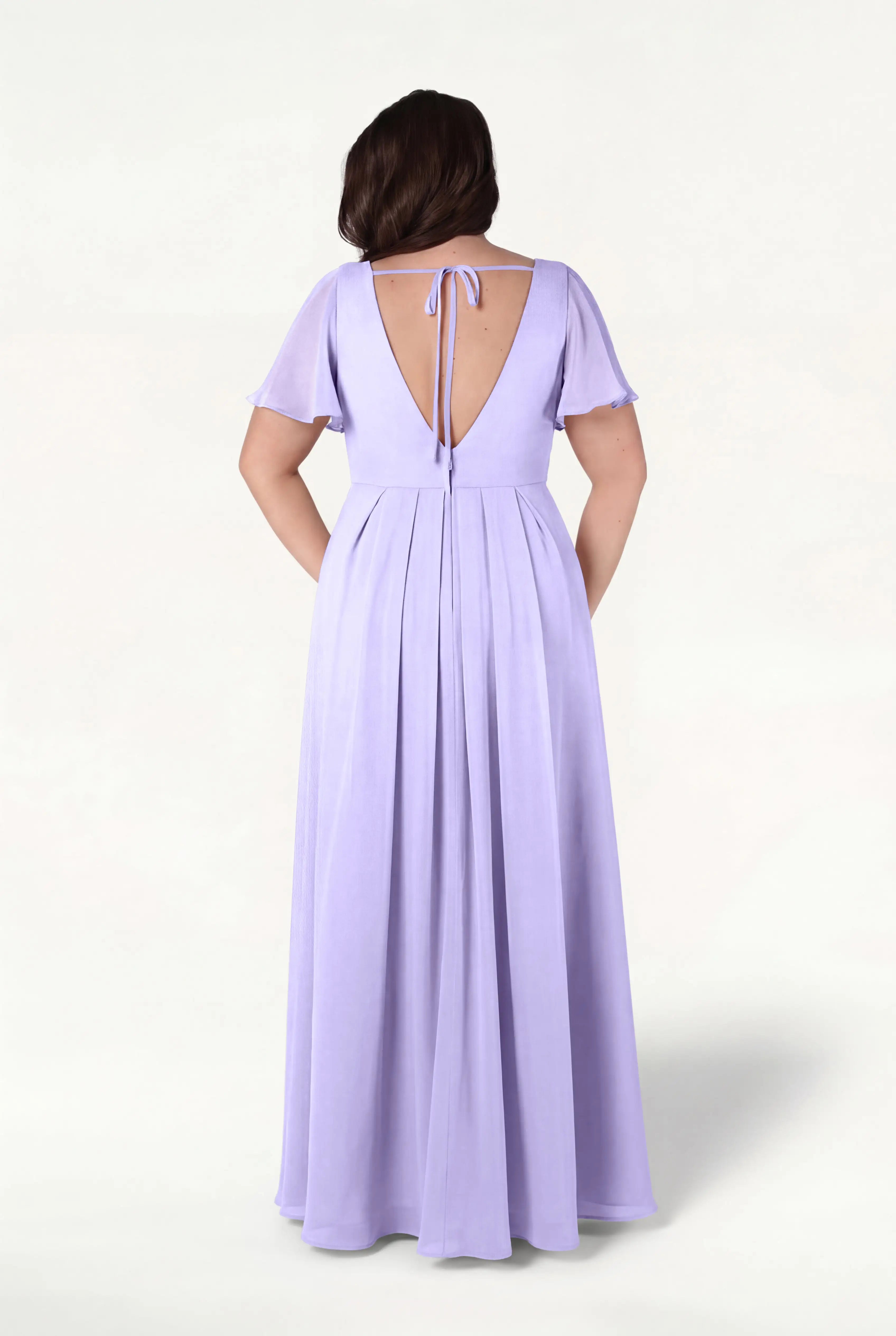 A woman stands with her back to the camera in a TH&TH Phoebe Bridesmaid Dress in Lilac Mist Chiffon, featuring a floor-length silhouette, short flutter sleeves, and a deep V-back tied with a bow—an effortlessly elegant bump-friendly choice.
