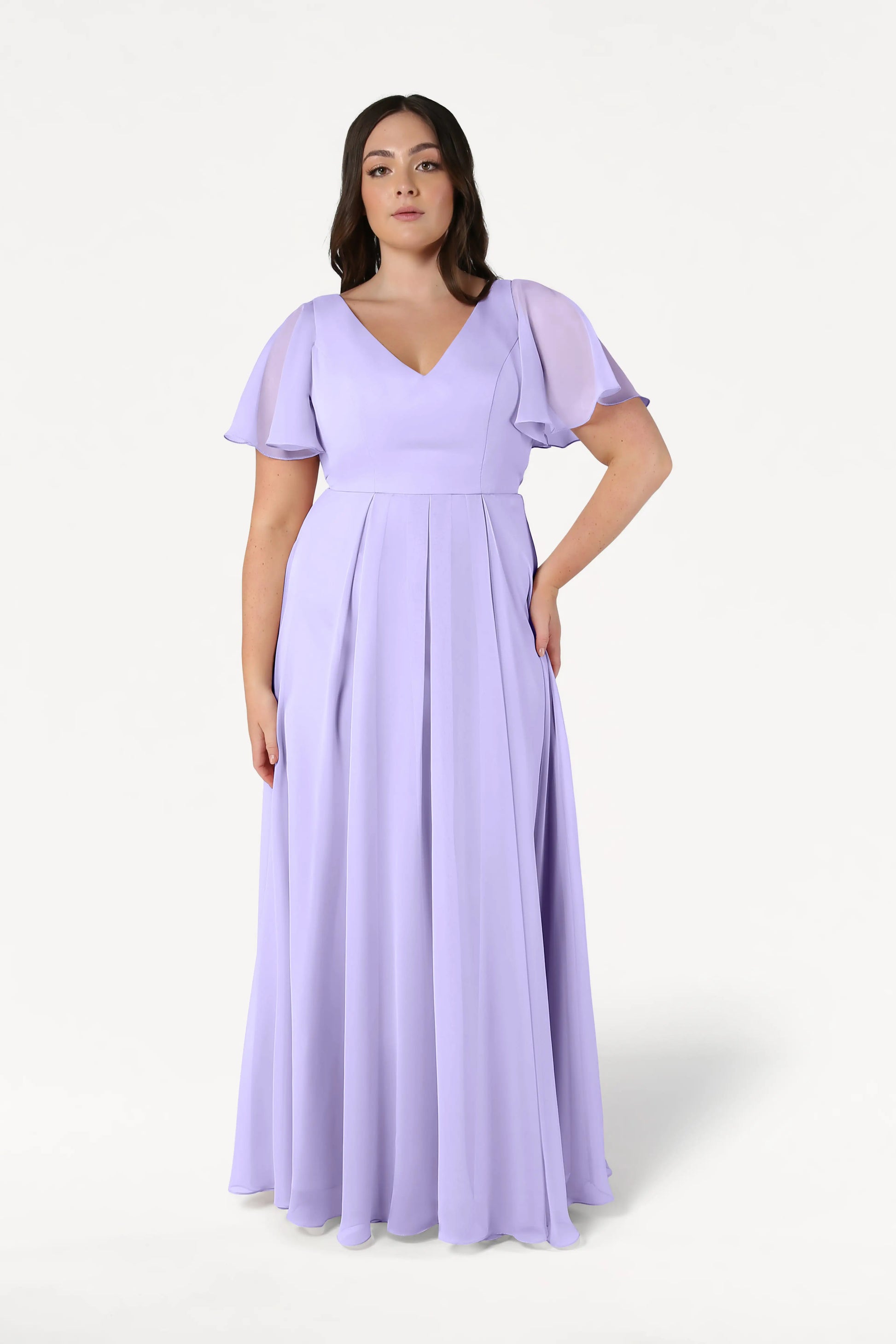 A woman wears the TH&TH Phoebe Bridesmaid Dress in Lilac Mist chiffon, featuring short flutter sleeves and a V-neckline, standing against a plain white background. This elegant gown is ideal for special occasions.