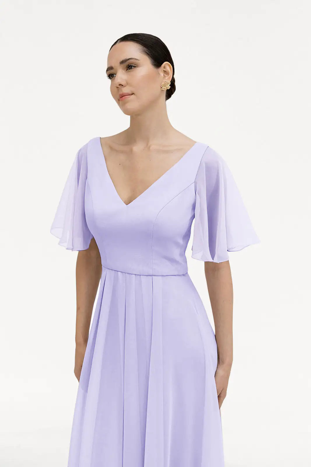 A woman with dark hair in a low bun wears the TH&TH Phoebe Bridesmaid Dress in Lilac Mist Chiffon, featuring flutter sleeves and a V-neckline, standing against a plain white background.