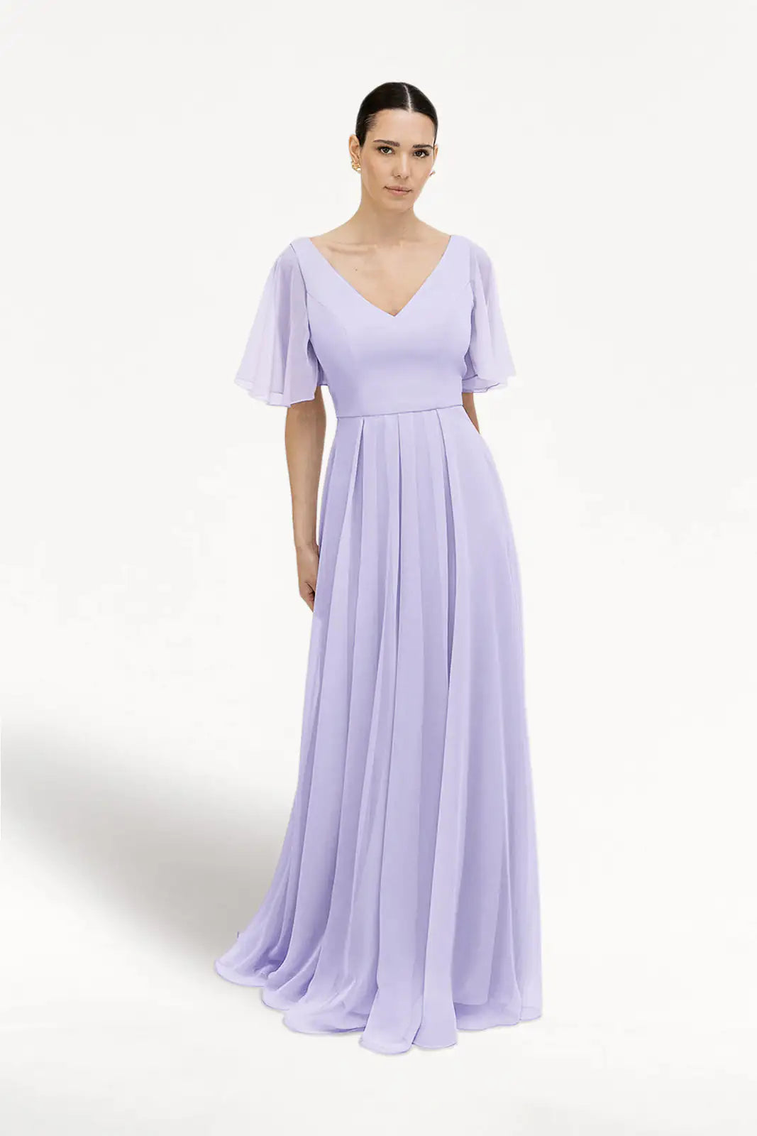 A woman wears the TH&TH Phoebe Bridesmaid Dress in Lilac Mist chiffon, featuring a floor-length, pleated skirt, short flutter sleeves, V-neckline, and fitted waist. The background is plain white.