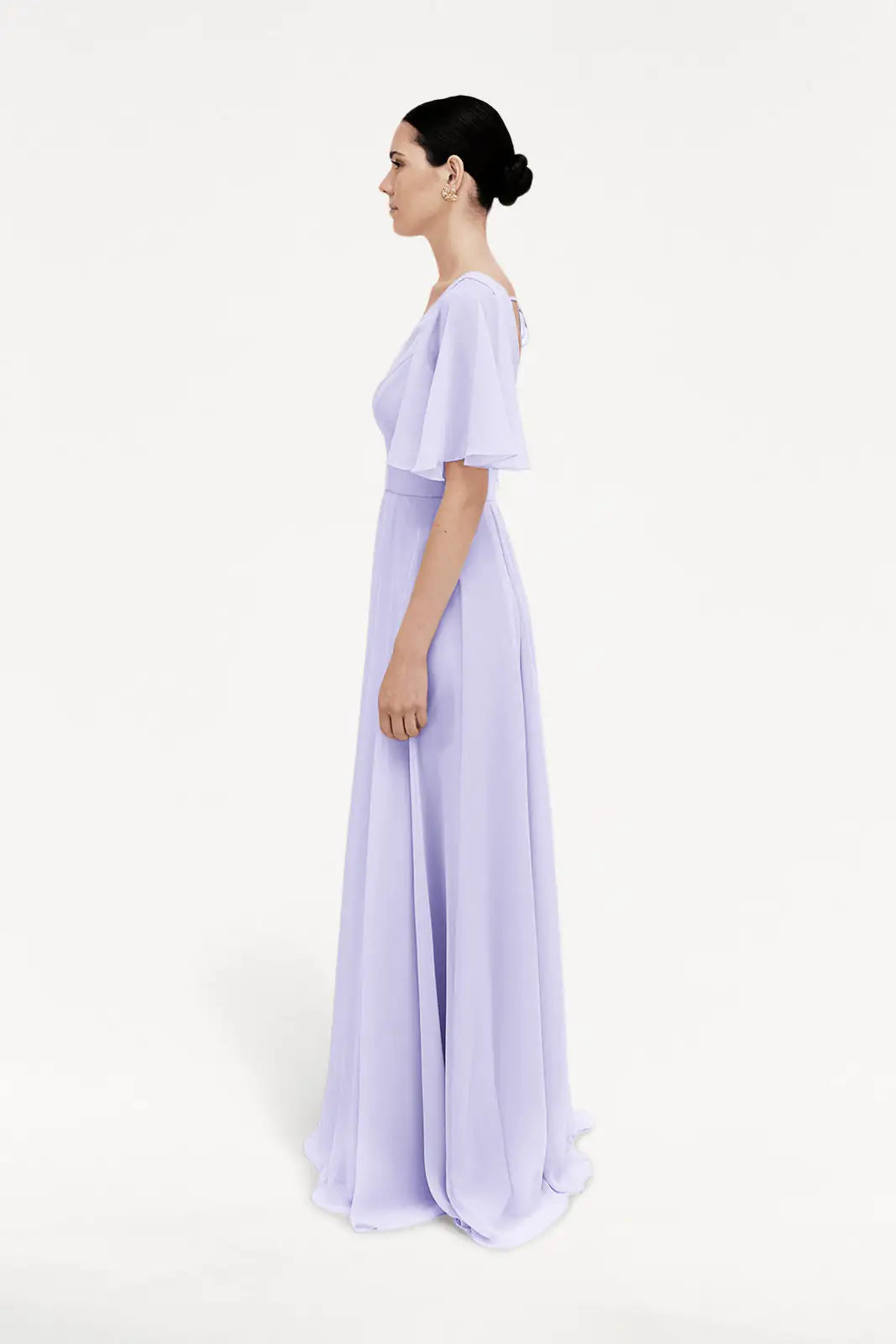 A woman with dark hair in a low bun stands in profile wearing the TH&TH Phoebe Bridesmaid Dress in Lilac Mist chiffon, a floor-length gown with flutter sleeves, perfect for bridesmaids, set against a plain white background.