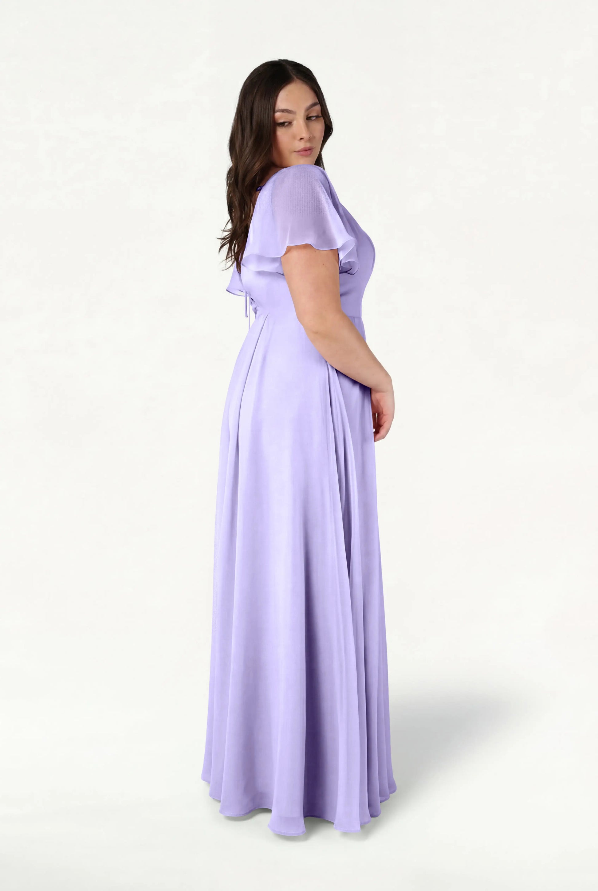 A woman with long brown hair stands sideways, wearing the TH&TH Phoebe Bridesmaid Dress in Lilac Mist Chiffon, a short-sleeved flowing gown, set against a light background.