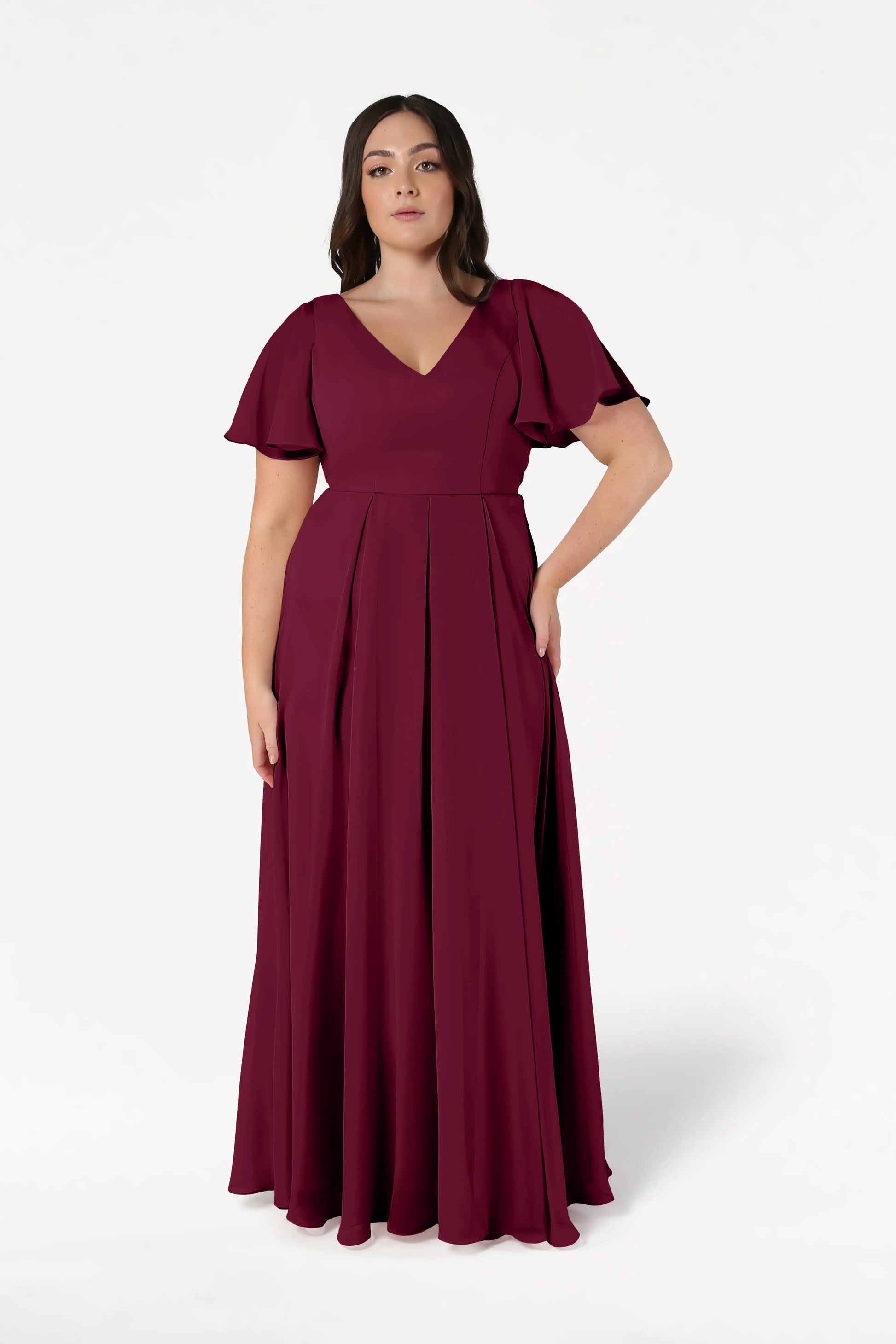 A woman stands against a white background wearing the TH&TH Phoebe Bridesmaid Dress in merlot chiffon, a floor-length, size-inclusive gown with short sleeves and a V-neckline.