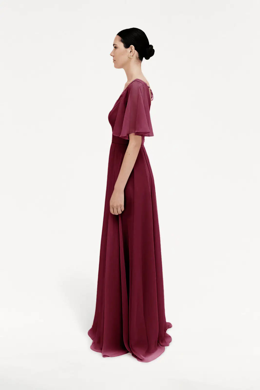 A woman in profile wears the TH&TH Phoebe Bridesmaid Dress in Merlot Chiffon, featuring short flutter sleeves and a flowing floor-length skirt. Her dark hair is styled in a low bun, making this gown ideal for bridesmaids.