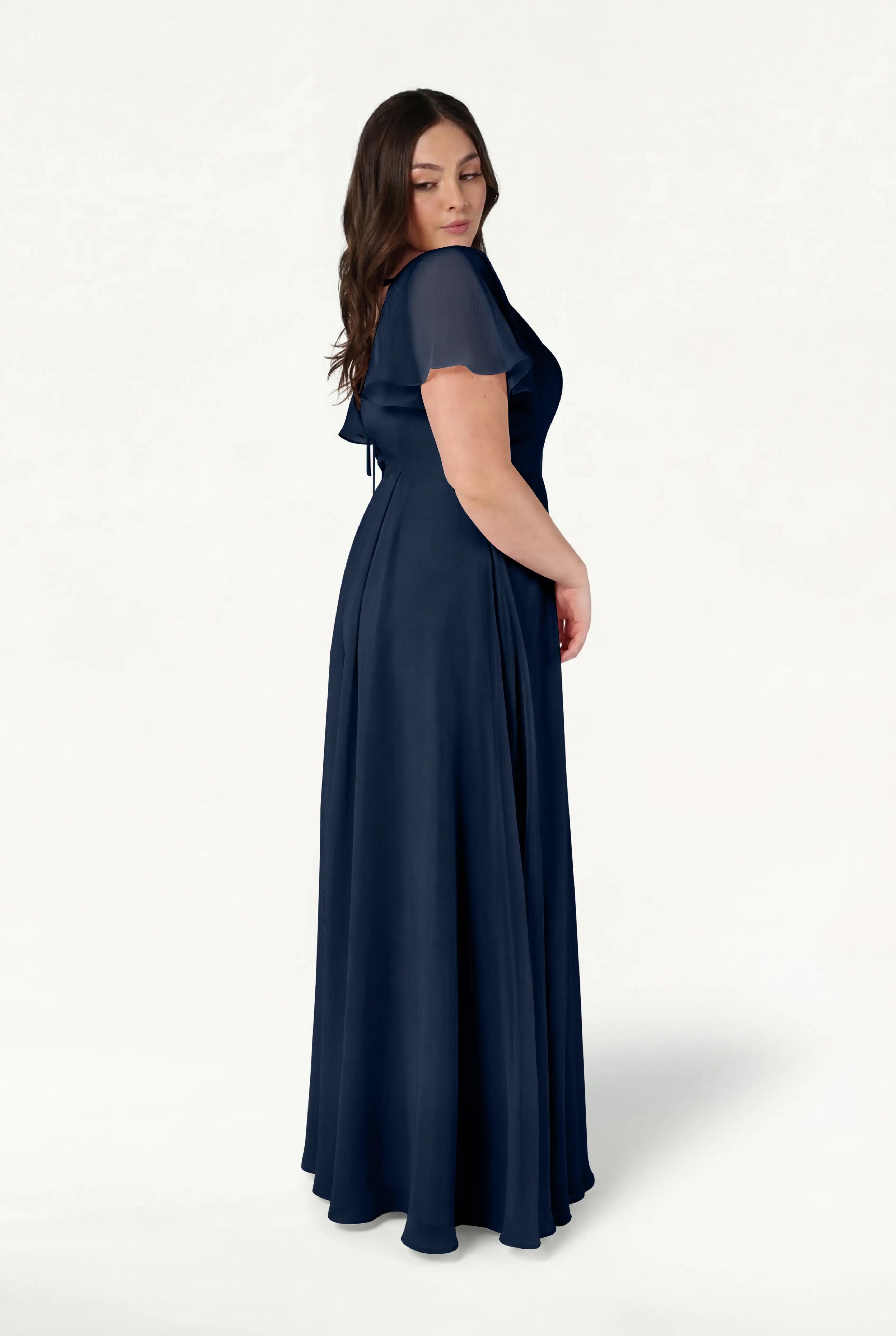A woman with long brown hair stands in profile against a white background wearing the TH&TH Phoebe Bridesmaid Dress in navy chiffon, featuring short sleeves and a flowing silhouette.