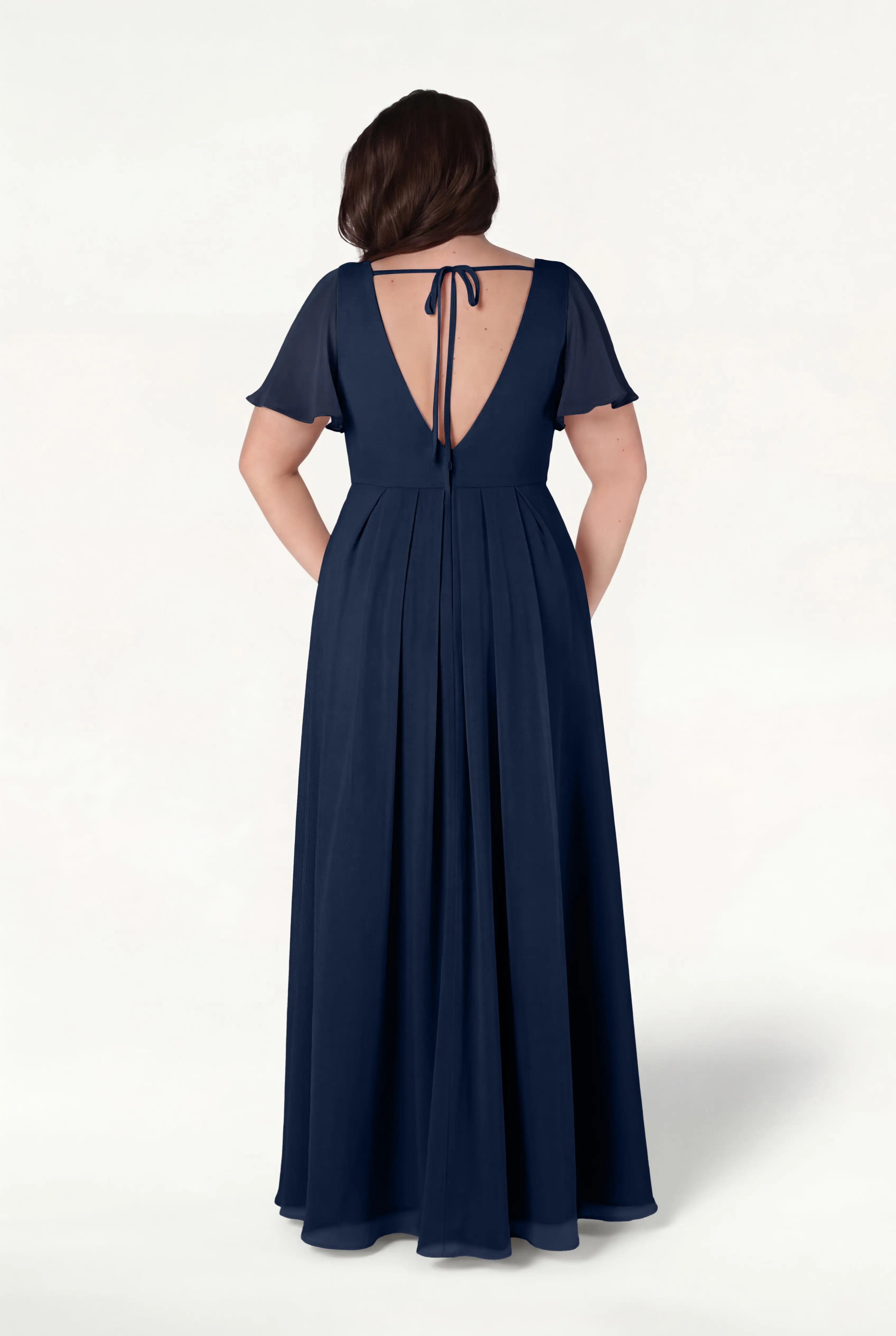 A woman is seen from the back in the TH&TH Phoebe Bridesmaid Dress in Navy Chiffon—a floor-length, short-sleeve gown featuring a V-shaped open back with a tie and a flowing, bump-friendly skirt.