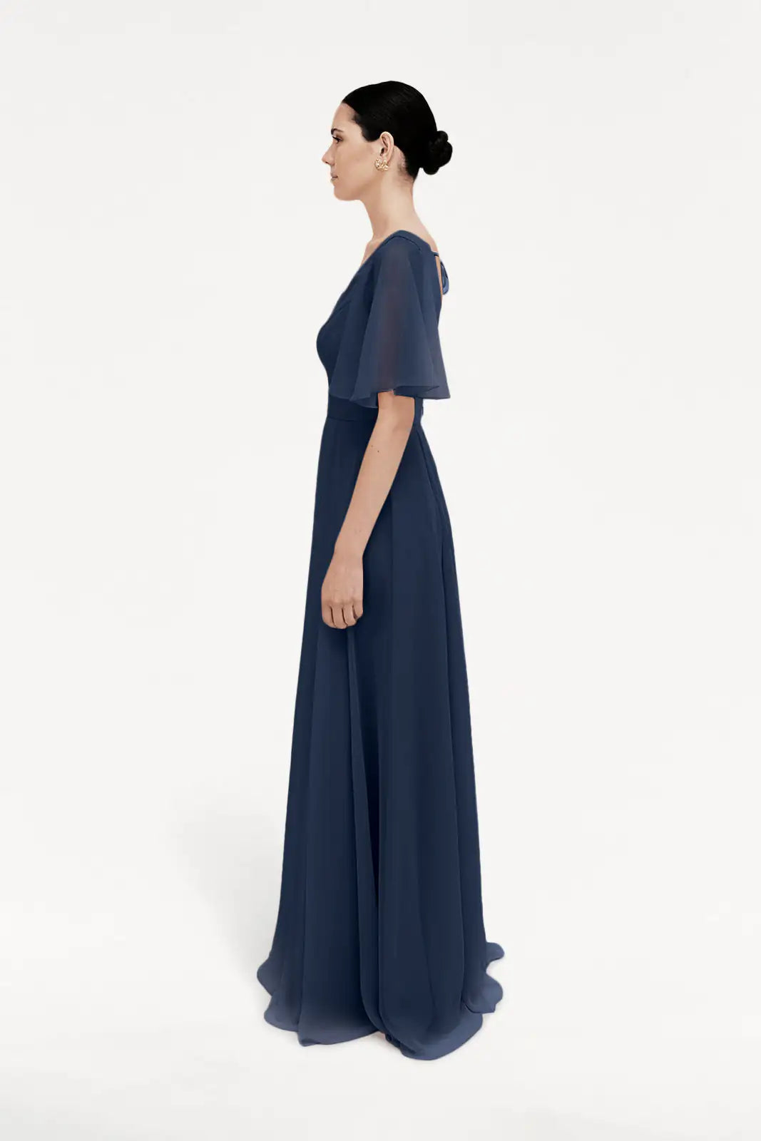 A woman in profile wears the TH&TH Phoebe Bridesmaid Dress in navy chiffon, a floor-length, bump-friendly gown with a fitted waist and sheer overlay, set against a plain white background.