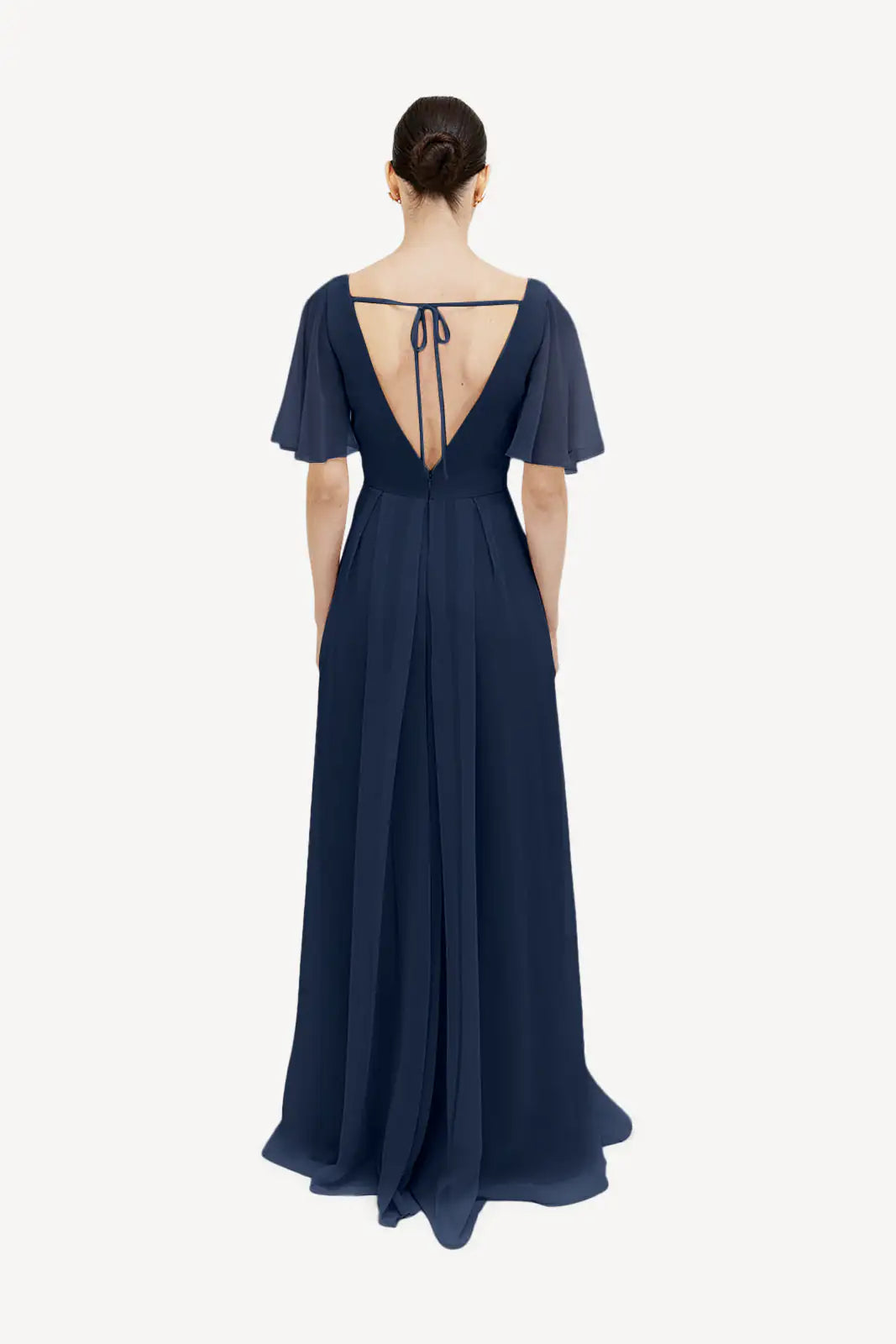 A woman in the TH&TH Phoebe Bridesmaid Dress in navy chiffon, featuring short flutter sleeves, a deep V-back with tie straps, and a flowing skirt, stands facing away with her hair in a bun.
