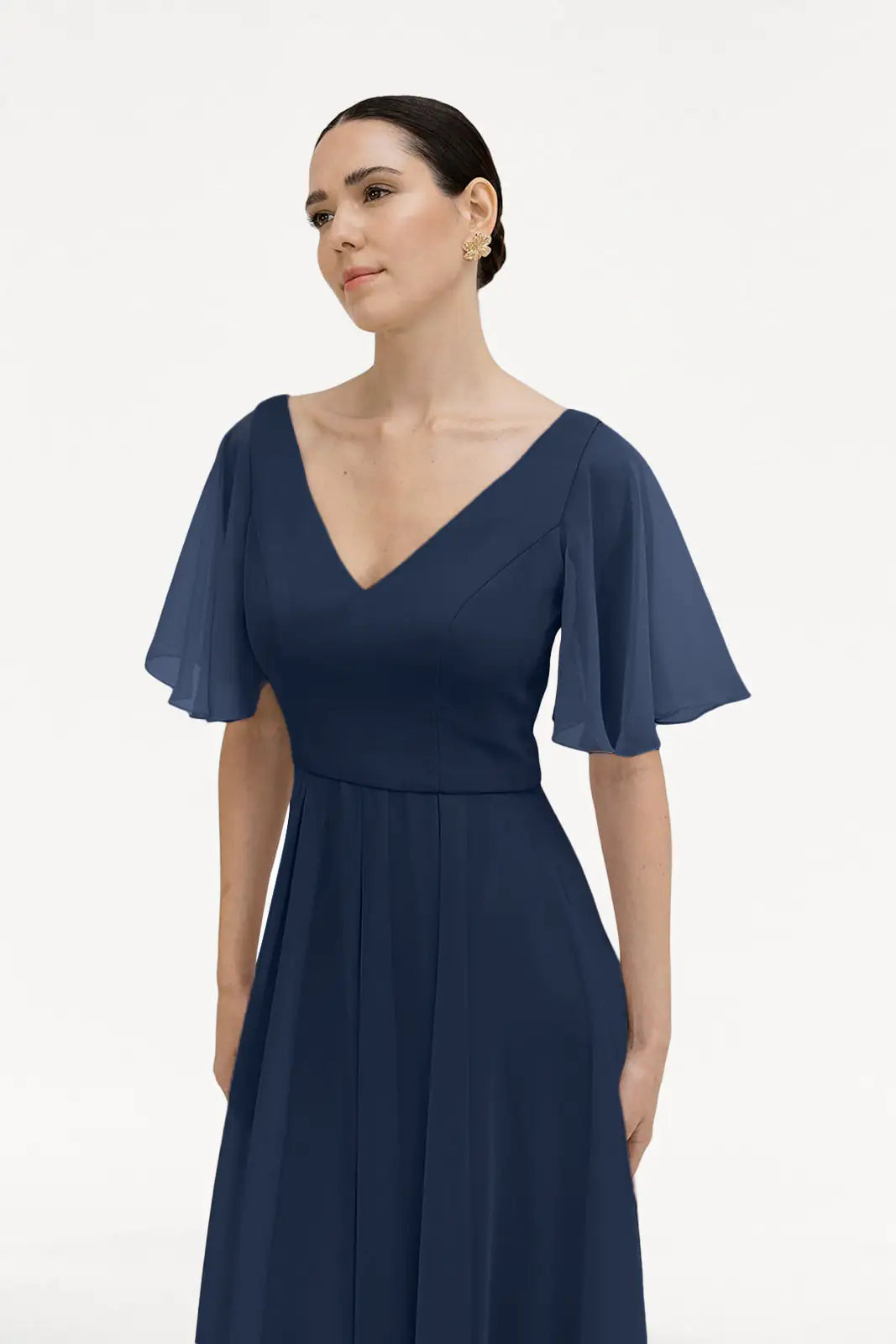 A woman in the TH&TH Phoebe Bridesmaid Dress in navy chiffon, featuring flutter sleeves and a deep V-neckline, stands against a white background with a neutral expression. This chic gown is also bump-friendly for expectant mothers.