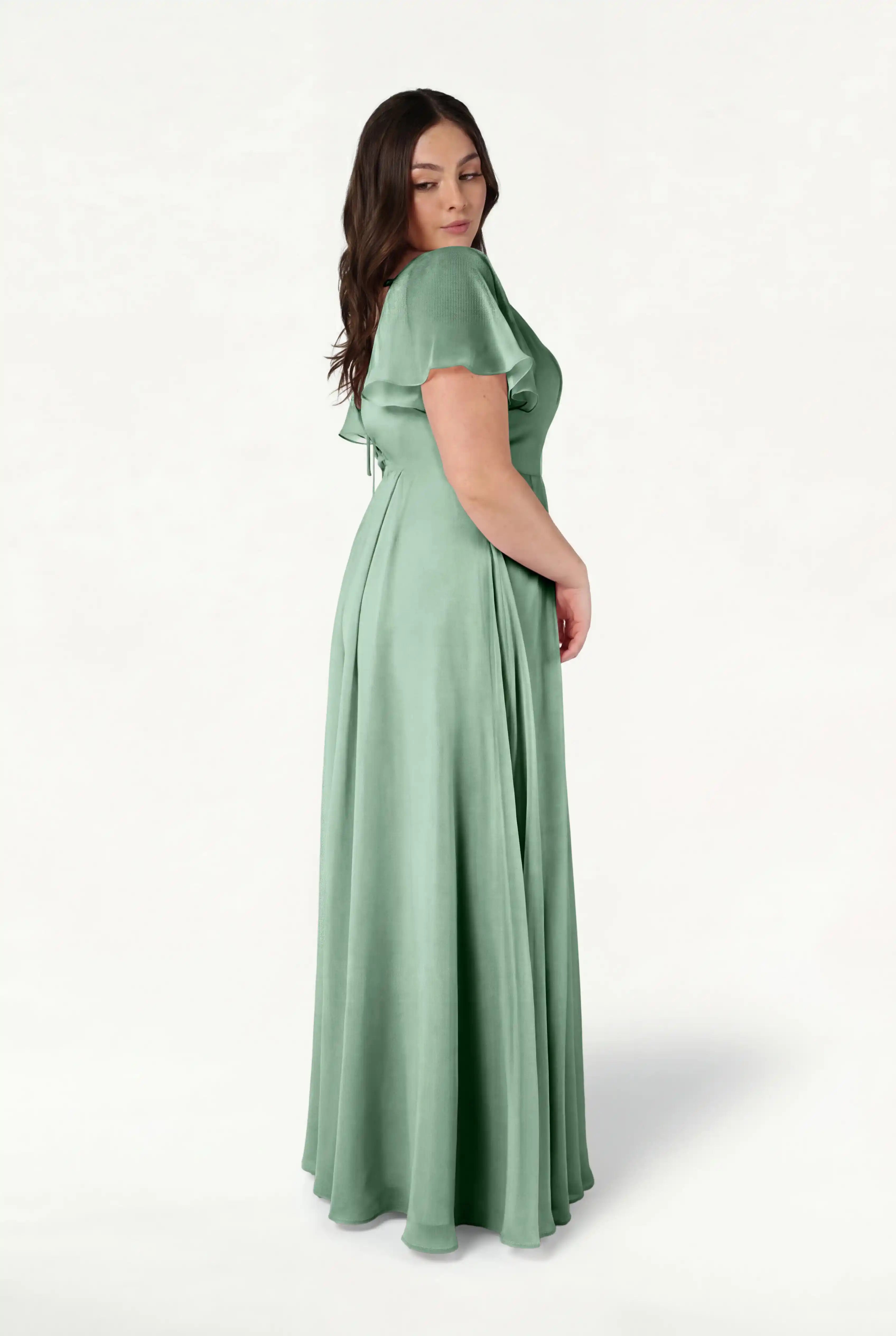 A woman with long brown hair stands in profile against a plain white background, wearing the TH&TH Phoebe Bridesmaid Dress in sage green chiffon—a bump-friendly, floor-length gown with short sleeves and soft, flowing fabric.