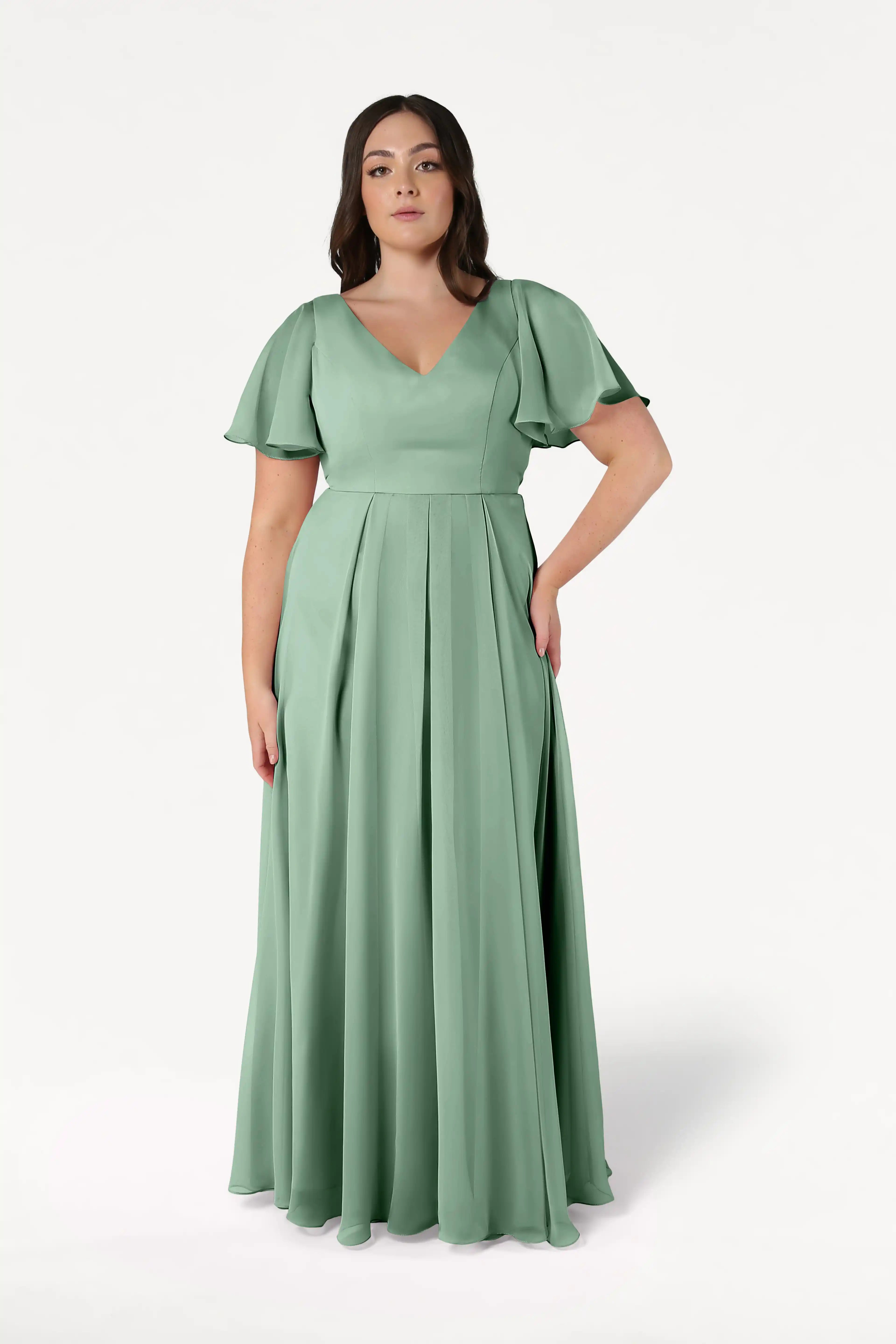 A woman with long, dark, wavy hair stands against a white background wearing the TH&TH Phoebe Bridesmaid Dress in sage green chiffon, featuring a floor-length skirt, short flutter sleeves, V-neckline, and fitted waist—an elegant bridesmaid style.