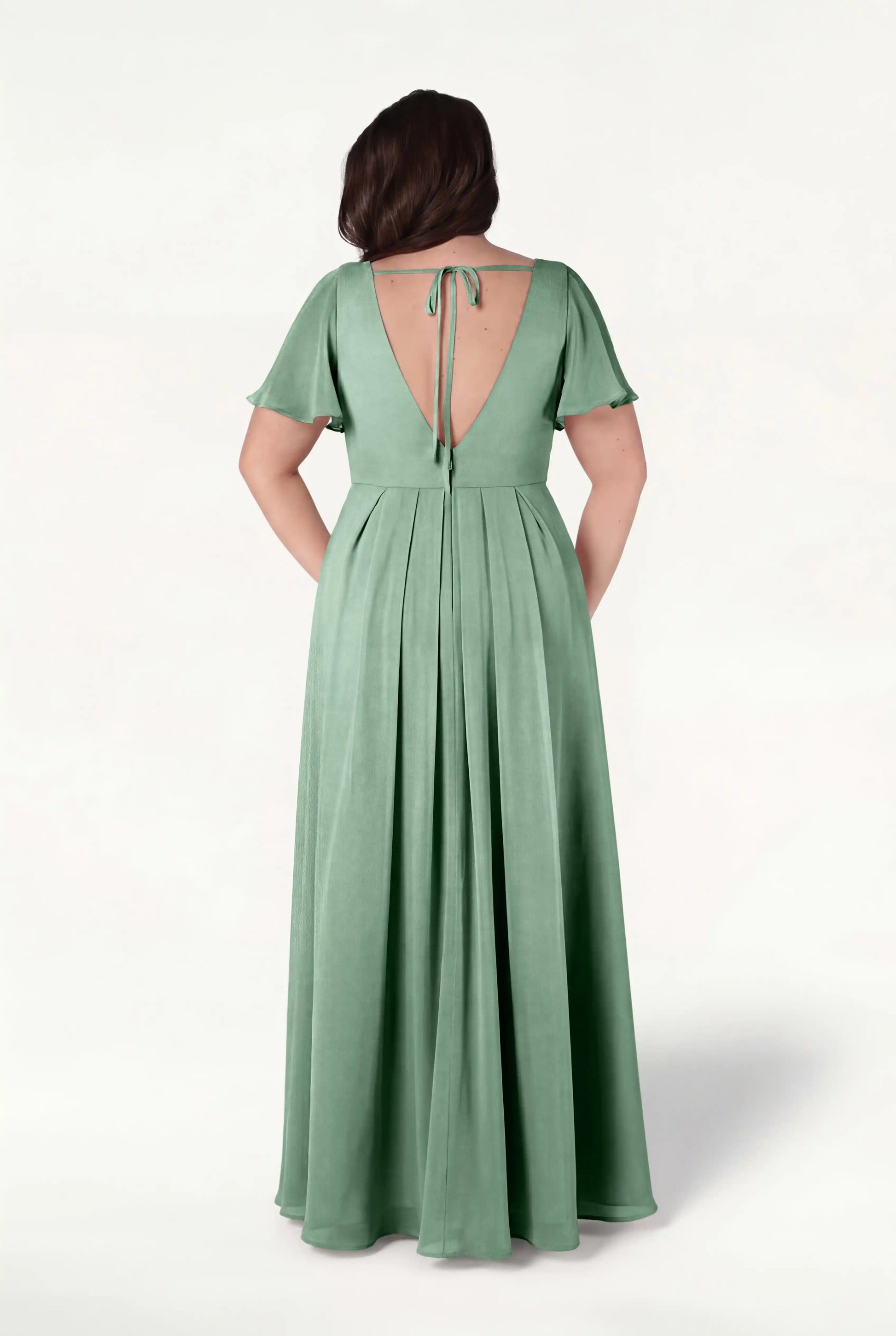 A woman models the TH&TH Phoebe Bridesmaid Dress in sage green chiffon, featuring short flutter sleeves, a deep V-back with a tie, soft pleats, and a flowing floor-length skirt for an elegant, bump-friendly look.