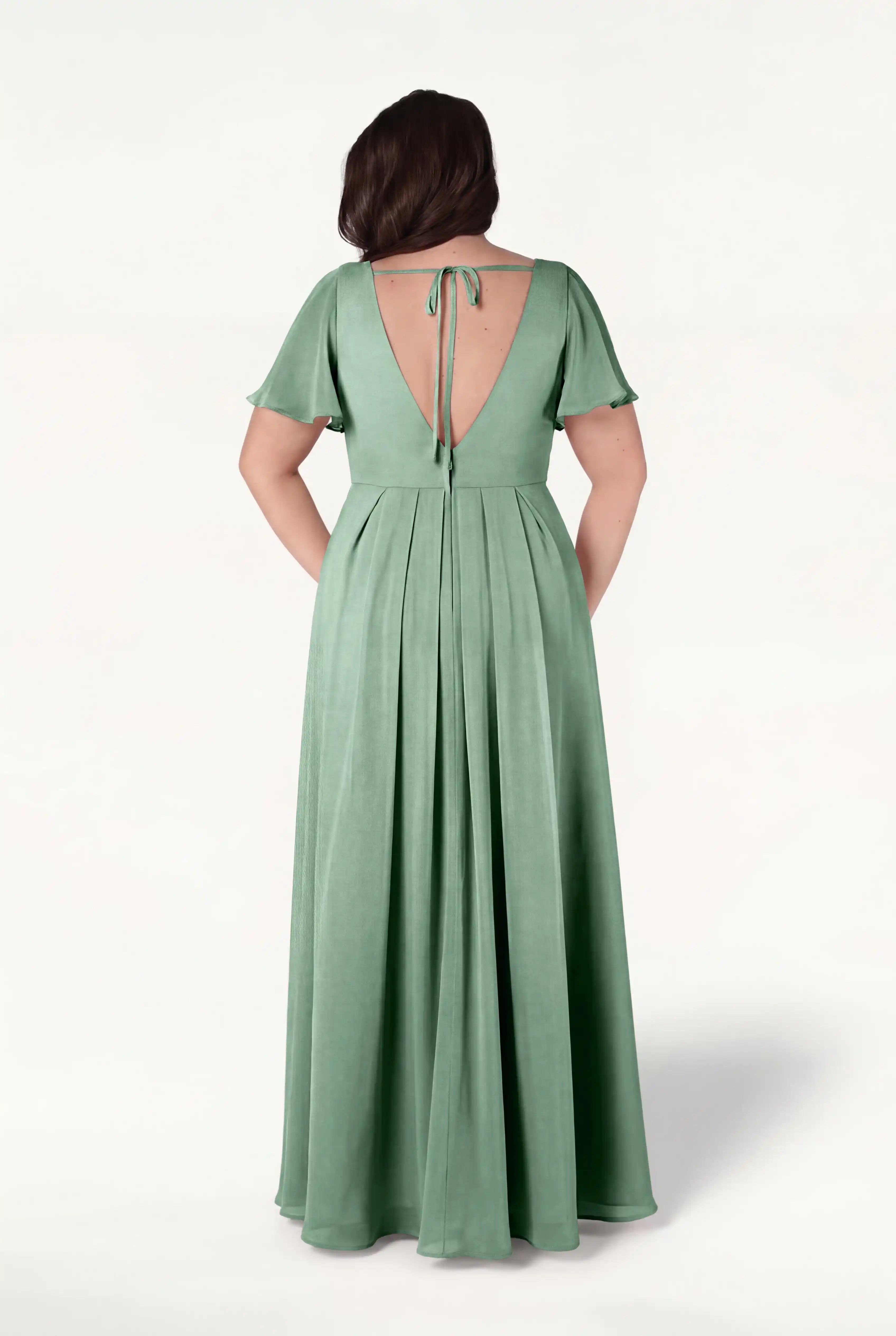A woman models the TH&TH Phoebe Bridesmaid Dress in sage green chiffon, featuring short flutter sleeves, a deep V-back with a tie, soft pleats, and a flowing floor-length skirt for an elegant, bump-friendly look.