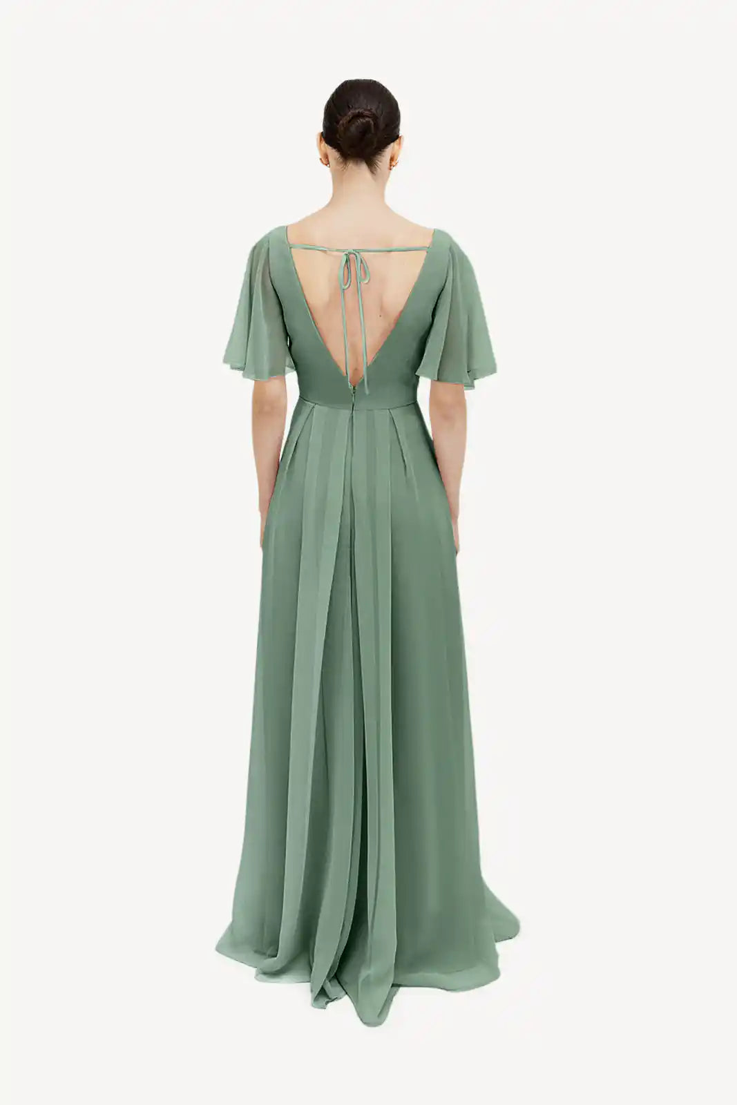 A woman stands with her back to the camera in the TH&TH Phoebe Bridesmaid Dress— a floor-length, sage green chiffon gown featuring short flutter sleeves, a deep V-back with tie straps, and a bump-friendly design.