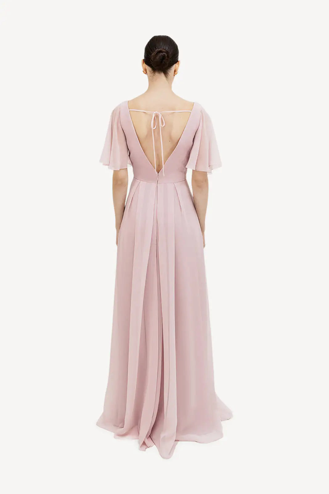 A woman stands facing away in the TH&TH Phoebe Bridesmaid Dress in Smoked Blush Chiffon, a floor-length gown with short flutter sleeves, a deep V-back, and back tie. Her hair is in a sleek bun against a white background.