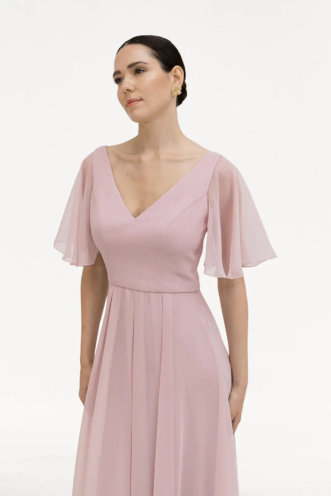 A woman models the Phoebe Bridesmaid Dress in Smoked Blush Chiffon by TH&TH, featuring bump-friendly chiffon and sheer flutter sleeves, standing against a plain white background with a soft, sideways gaze.