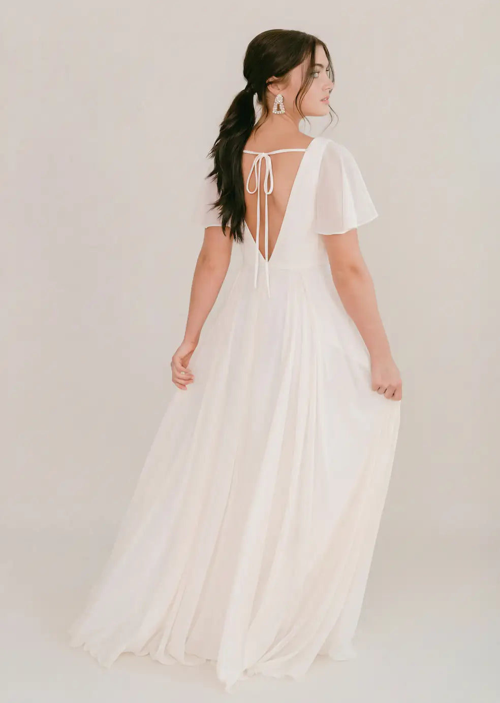 A woman models the TH&TH Phoebe Dress in Ivory Chiffon, a bump-friendly gown with flutter sleeves and a deep V-back tied with a bow. Her long dark hair falls in loose waves, complemented by statement earrings as she poses with her back to the camera.