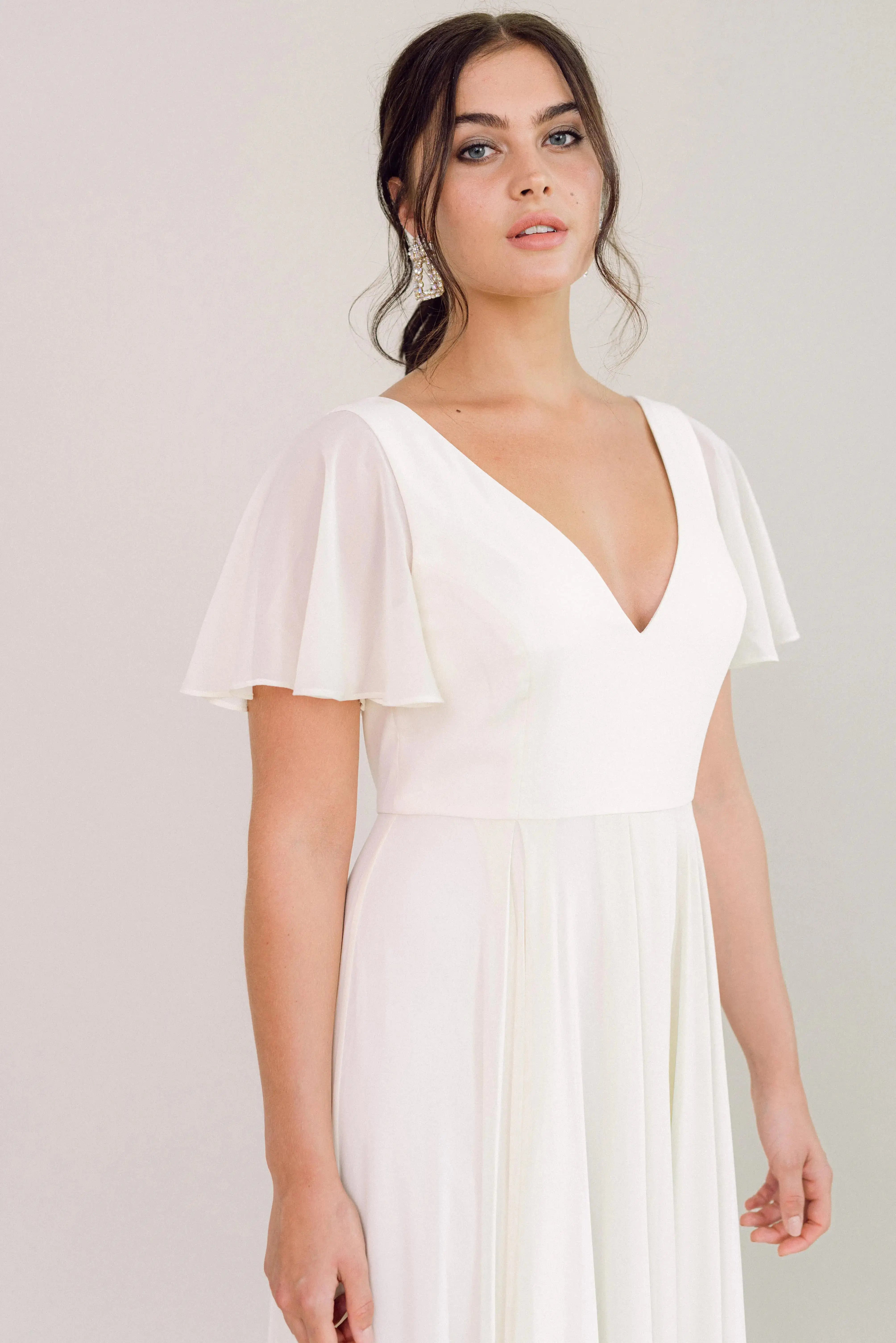 A woman wears the TH&TH Phoebe Dress in Ivory Chiffon, featuring flutter sleeves and a deep V-neckline—a chic, bump-friendly option for bridesmaids or special occasions.