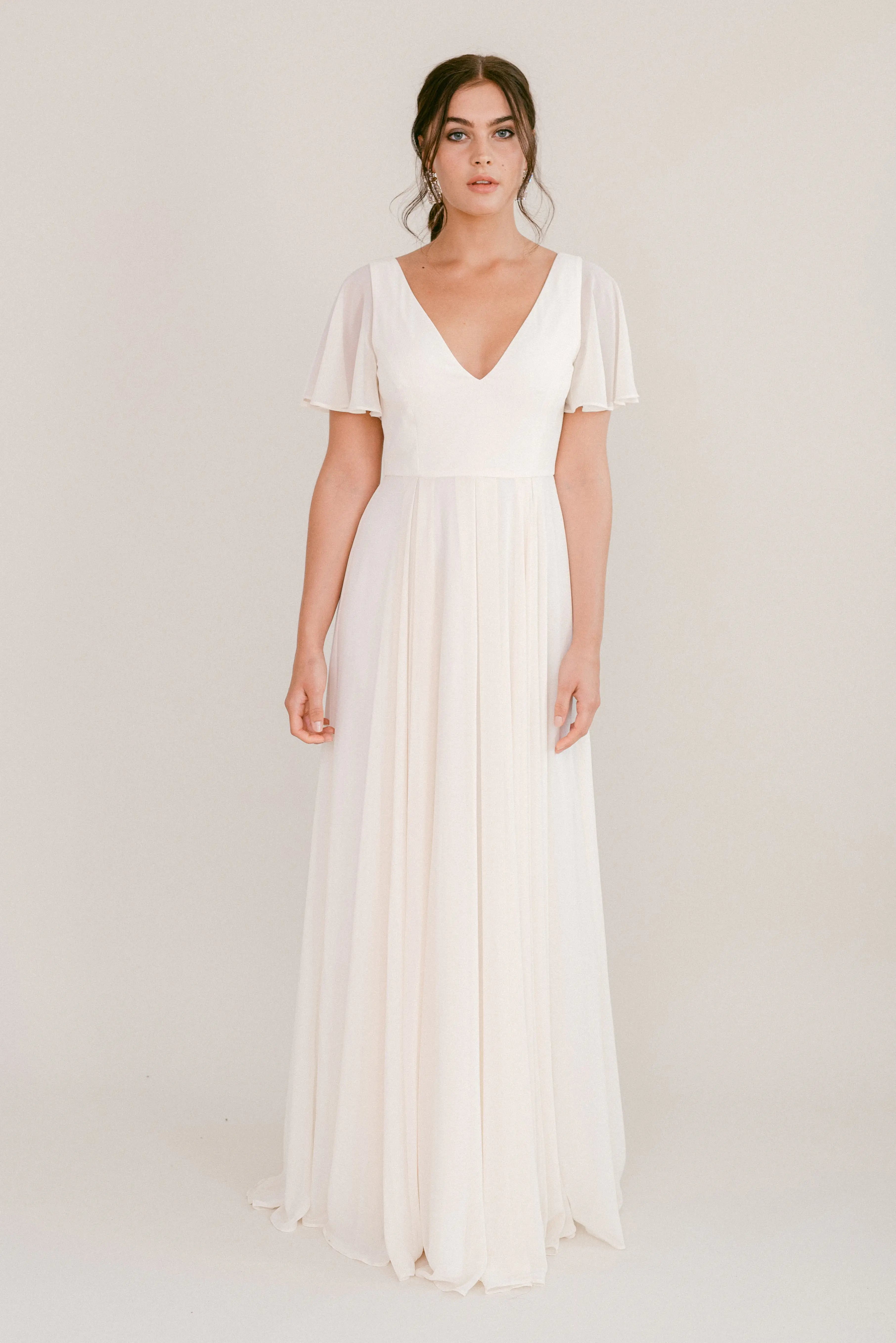 A woman with loose waves and a neutral expression wears the bump-friendly Phoebe Dress in Ivory Chiffon by TH&TH—a floor-length, short-sleeved gown featuring a deep V-neckline and flowing skirt, set against a plain background.