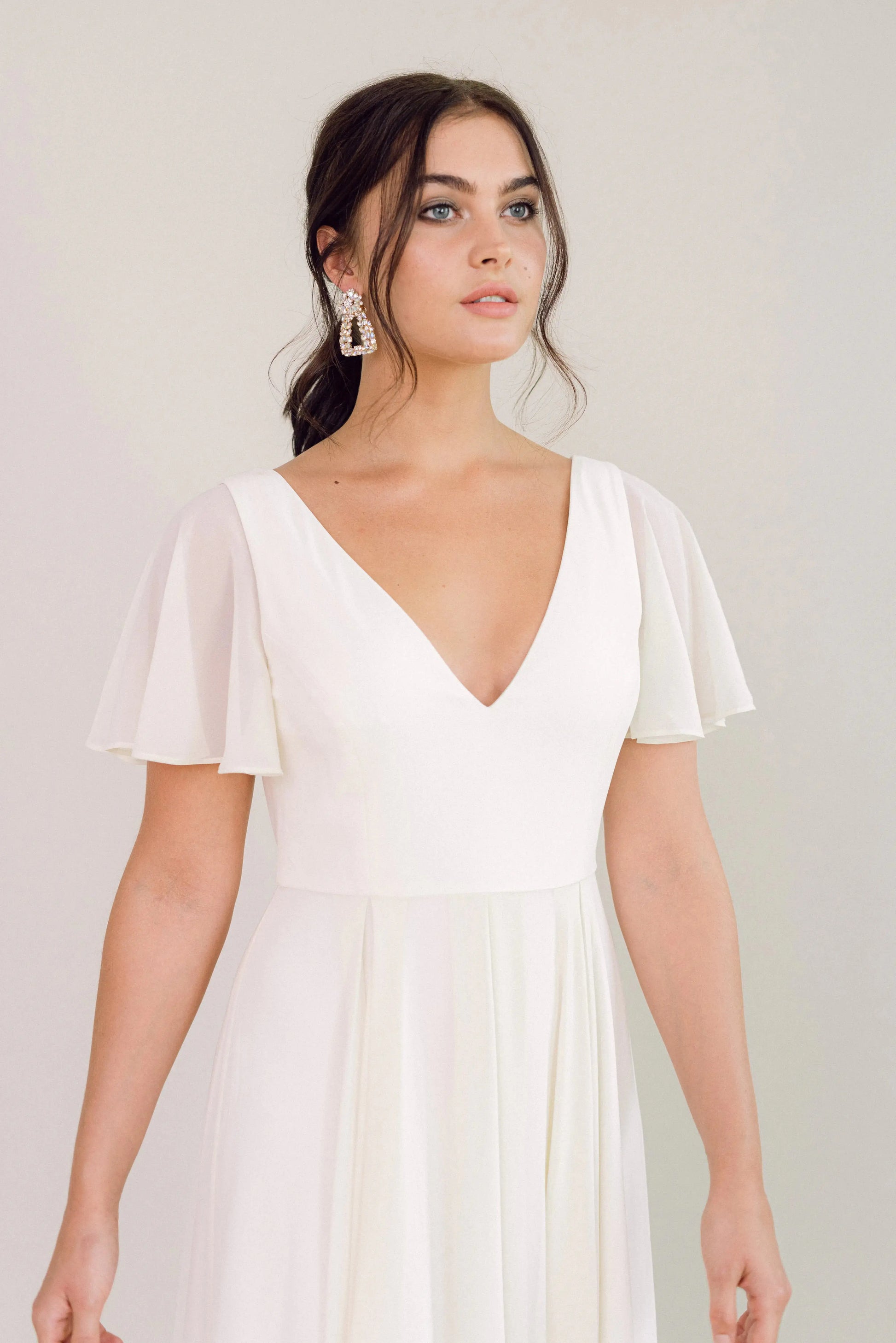 A woman models the TH&TH Phoebe Dress in Ivory Chiffon, featuring flutter sleeves and a deep V-neckline. Her dark hair is styled back and she wears statement earrings, creating an elegant bridesmaid look against a plain light background.