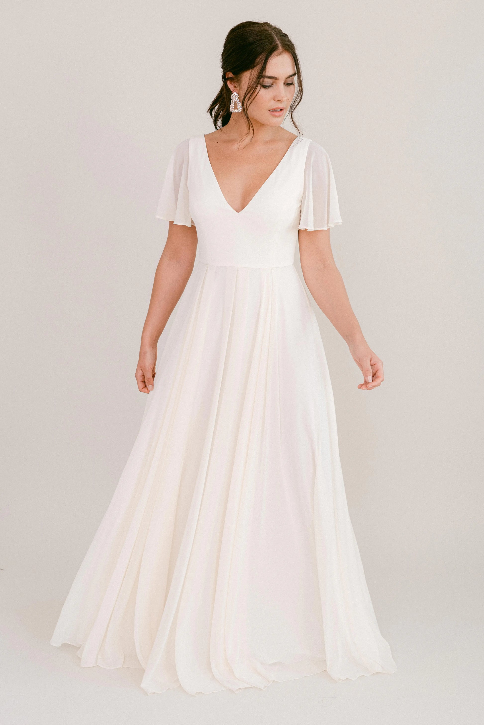 A woman with dark hair styled up wears the TH&TH Phoebe Dress in Ivory Chiffon, featuring a flowing, floor-length silhouette, short flutter sleeves, and a deep V neckline. She stands against a plain light background looking slightly downward.