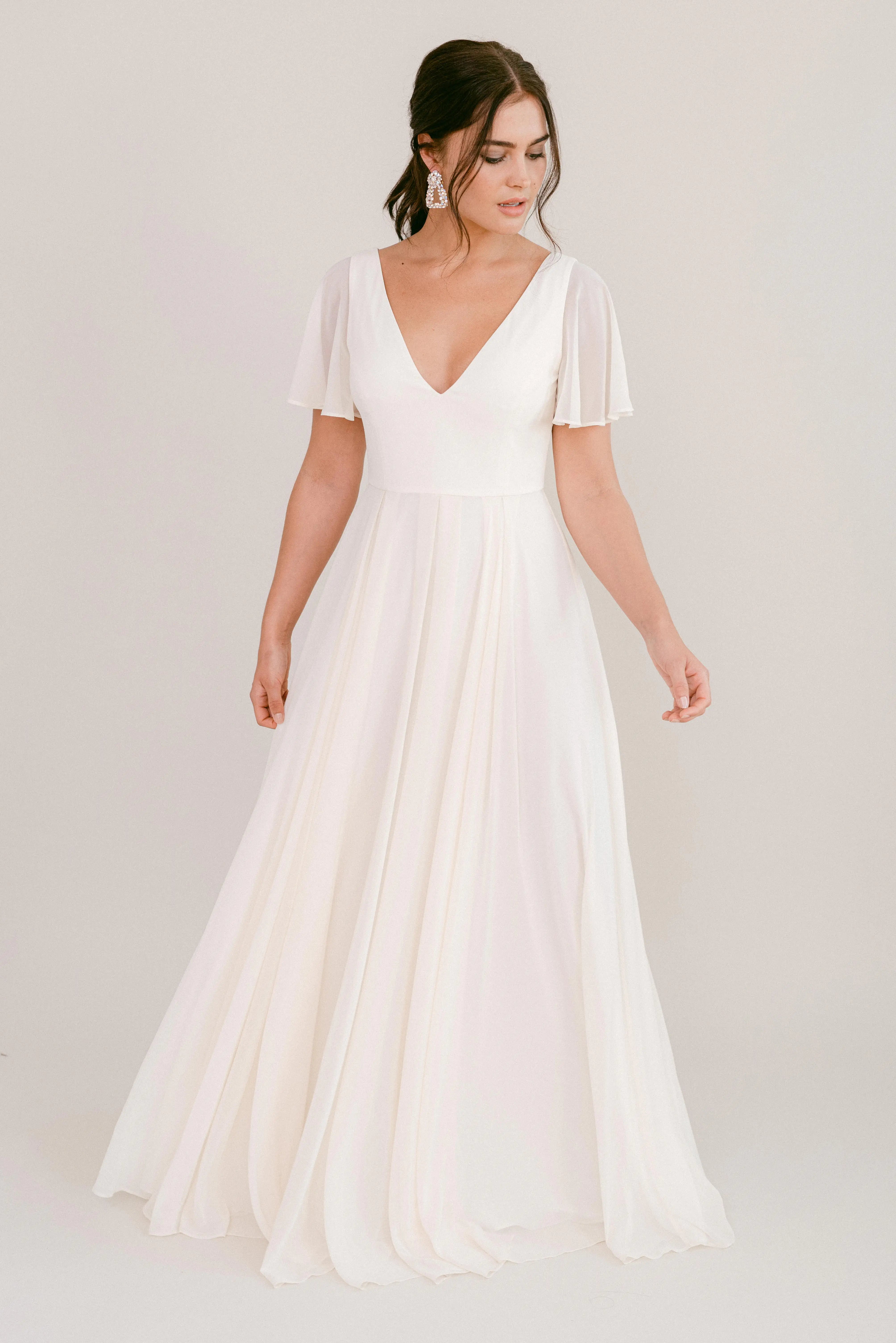 A woman with dark hair styled up wears the TH&TH Phoebe Dress in Ivory Chiffon, featuring a flowing, floor-length silhouette, short flutter sleeves, and a deep V neckline. She stands against a plain light background looking slightly downward.