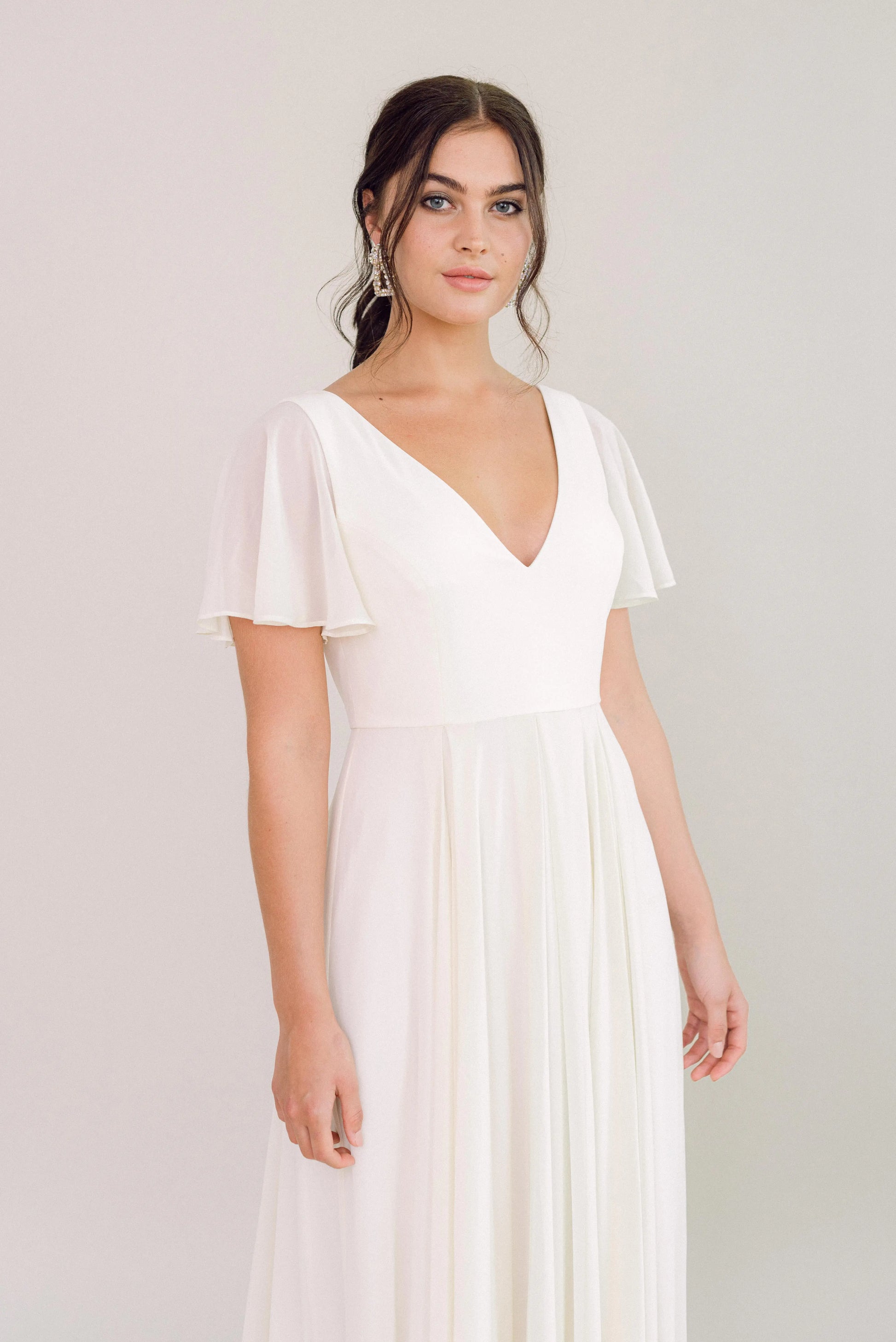 A woman models the TH&TH Phoebe Dress in Ivory Chiffon, a short-sleeved V-neck gown with a flowing skirt. She stands against a light backdrop with loose waves in her dark hair and a calm, neutral expression.