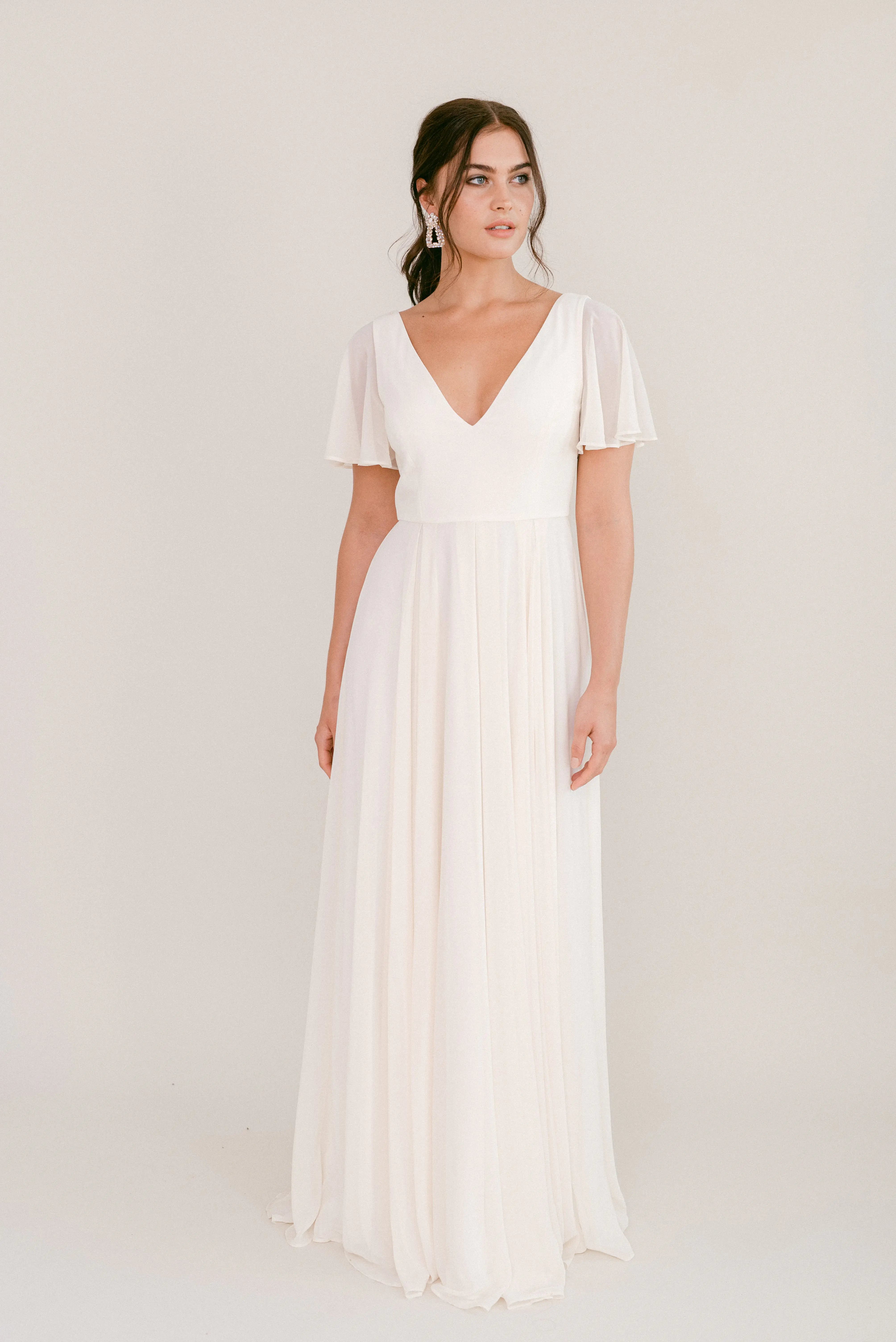 A woman stands against a plain background wearing the TH&TH Phoebe Dress in Ivory Chiffon, featuring short flutter sleeves and a deep V-neckline. Her dark hair is styled back as she gazes slightly to the side.