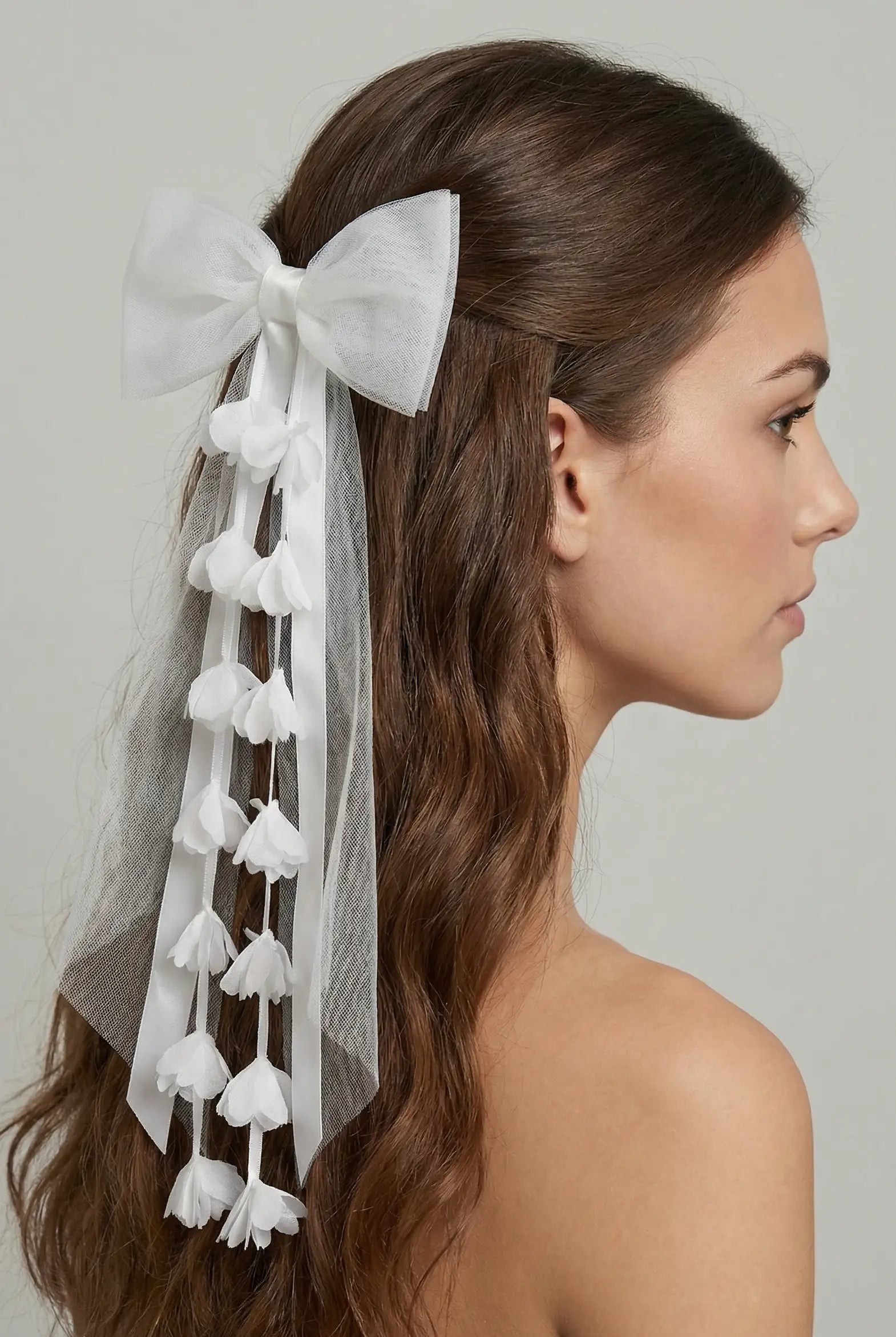 A woman with long, wavy brown hair wears the Posey Tulle Hair Bow by TH&TH—a large, elegant white bow styled at the back of her head, featuring trailing tulle ribbons and delicate white fabric flowers.