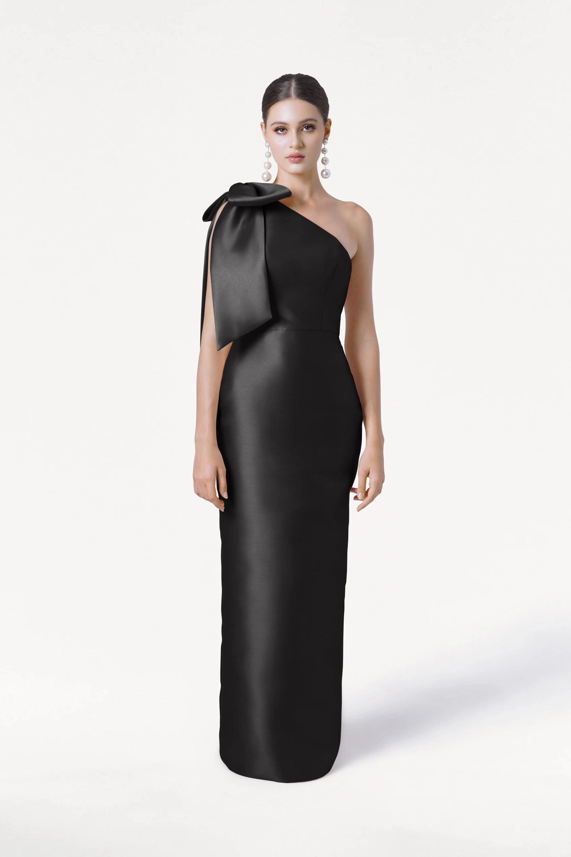 A woman models the TH&TH Ritz Bridesmaid Dress in Black Satin Twill, a sleek one-shoulder gown with a statement bow. Her hair is pulled back, and she wears statement earrings against a plain white background.