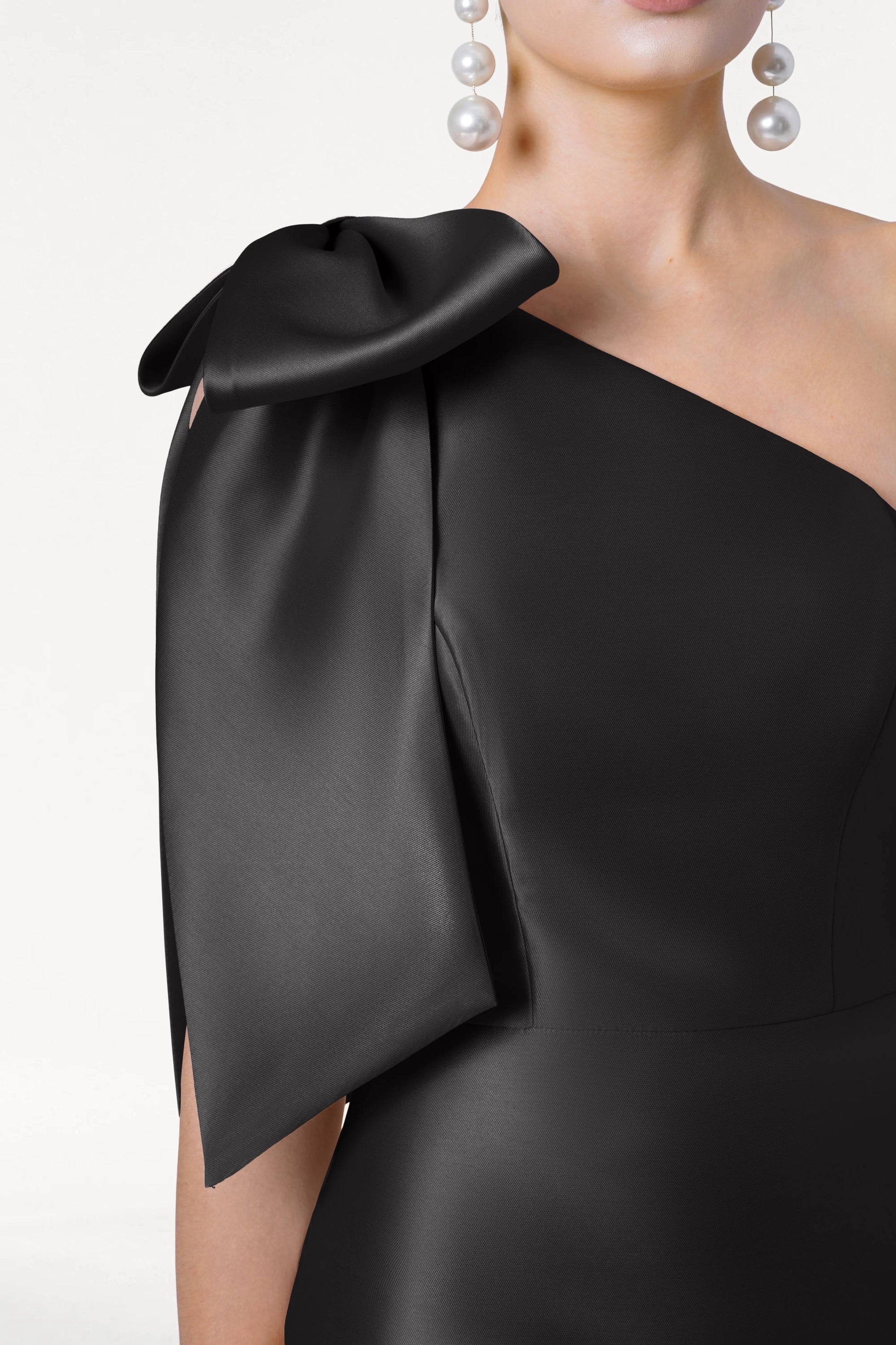 A woman models the TH&TH Ritz Bridesmaid Dress in Black Satin Twill—a one-shoulder gown with a statement shoulder bow—paired with long pearl drop earrings, shown from neck to waist on a plain white background.