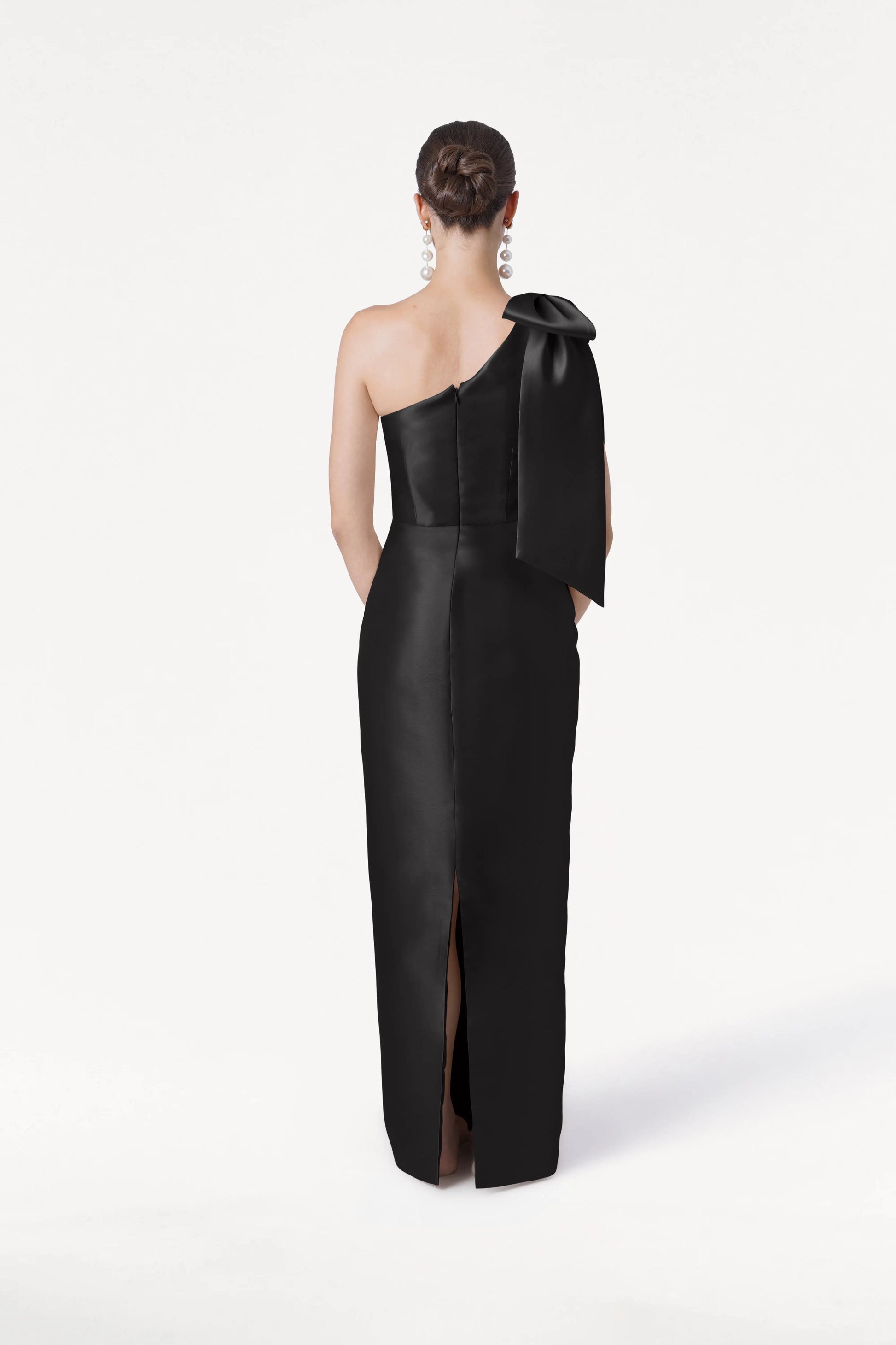 A woman with her hair in a bun models the TH&TH Ritz Bridesmaid Dress in Black Satin Twill, featuring a one-shoulder design with a statement bow and back slit. She wears large dangling earrings, standing against a plain white background.