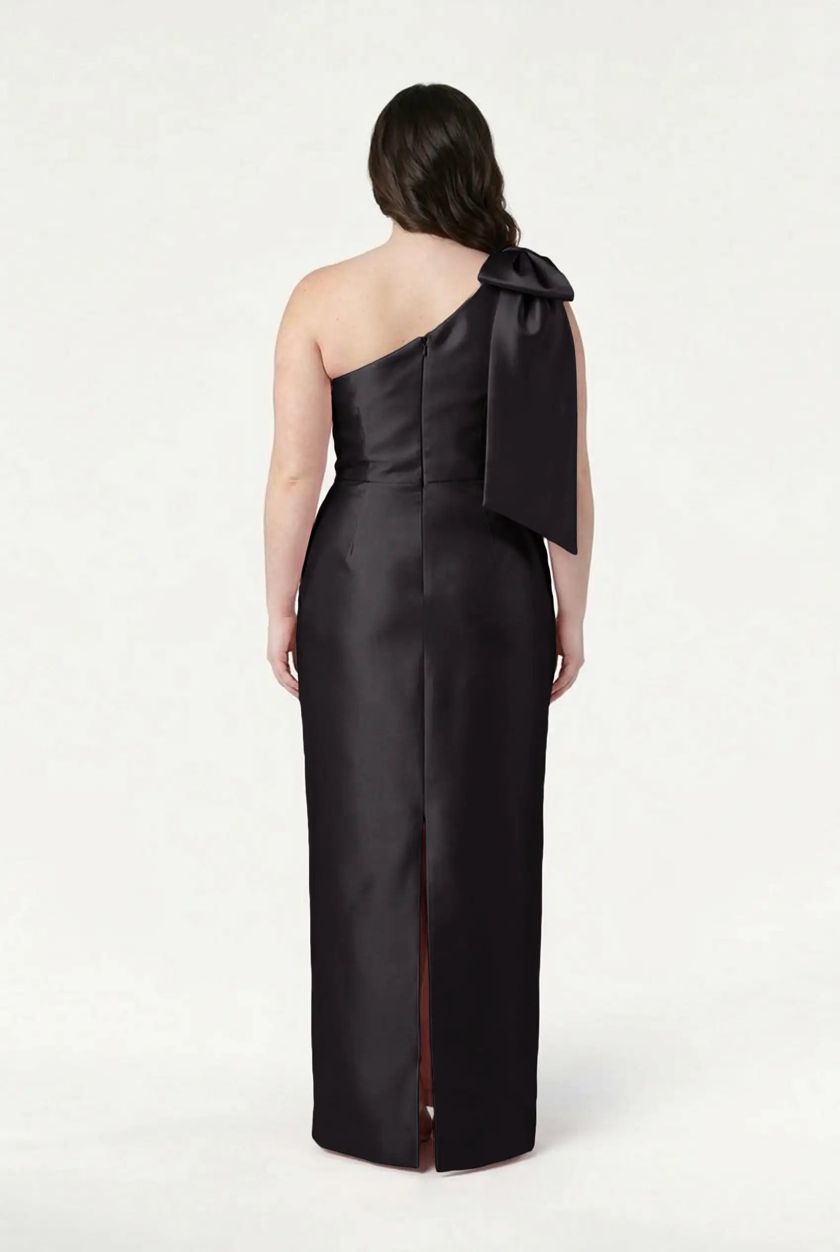 A woman with loose dark waves stands against a white background, wearing the TH&TH Ritz Bridesmaid Dress in Black Satin Twill—a one-shoulder gown featuring a statement bow and back slit.
