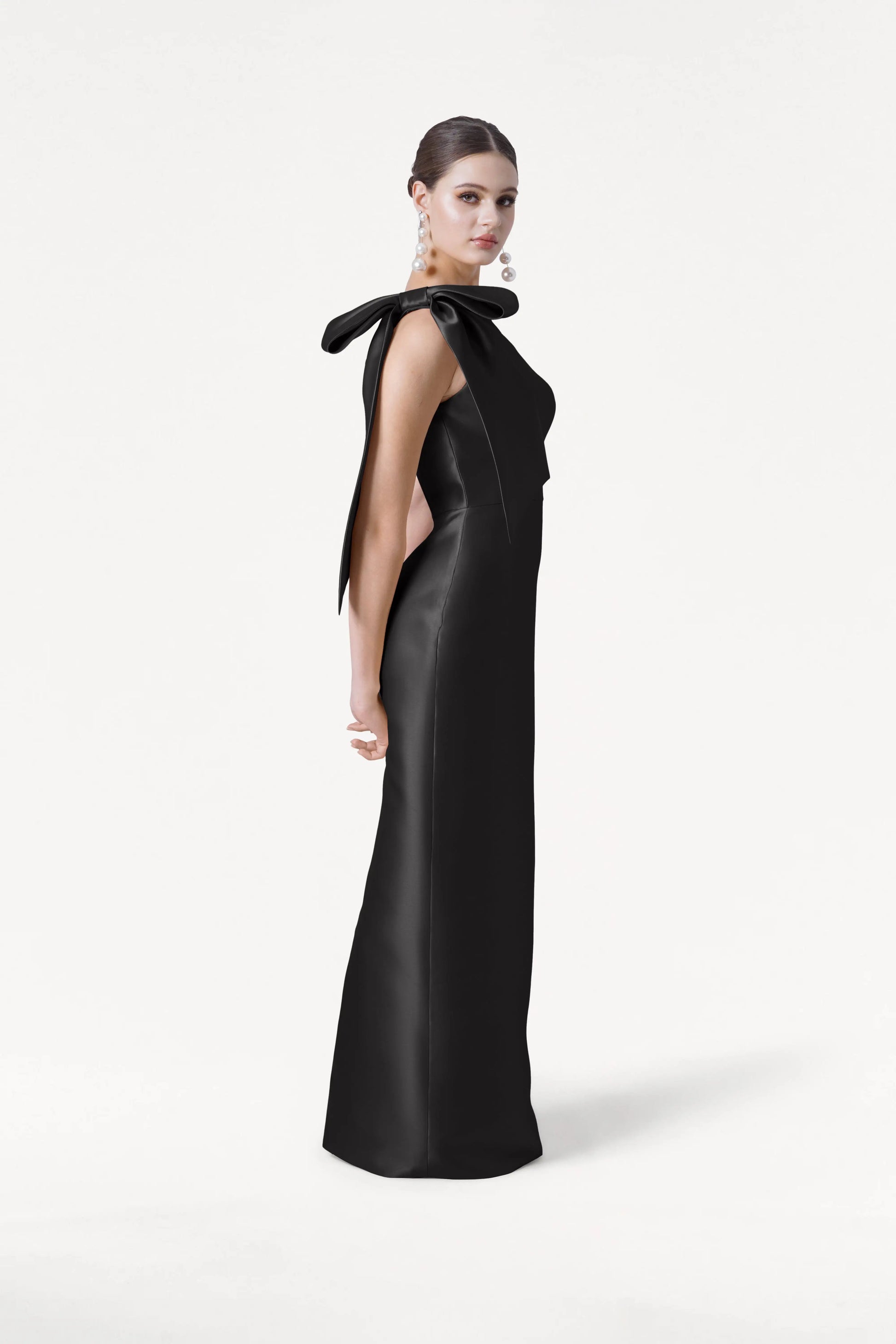 A woman wears the TH&TH Ritz Bridesmaid Dress in Black Satin Twill, featuring a floor-length silhouette, open back, and statement shoulder bow. She stands against a white background with her hair up and bold earrings.
