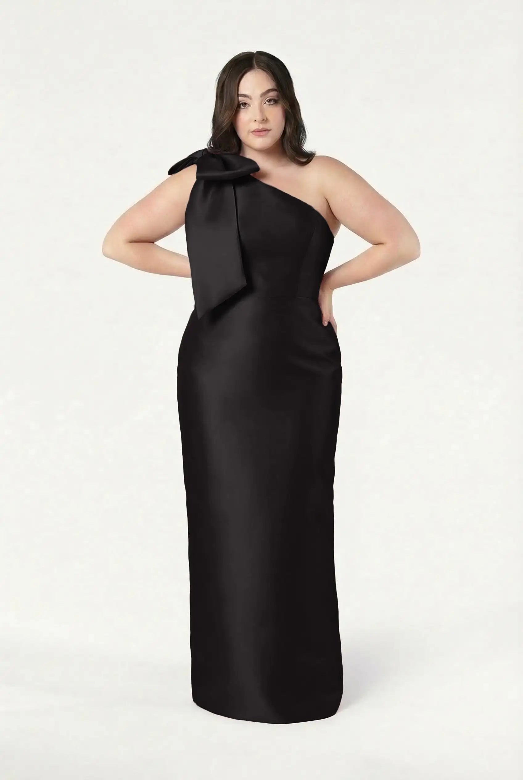 A woman confidently poses in the TH&TH Ritz Bridesmaid Dress, a floor-length black satin twill gown with a one-shoulder design and statement bow, standing out against a plain white background.