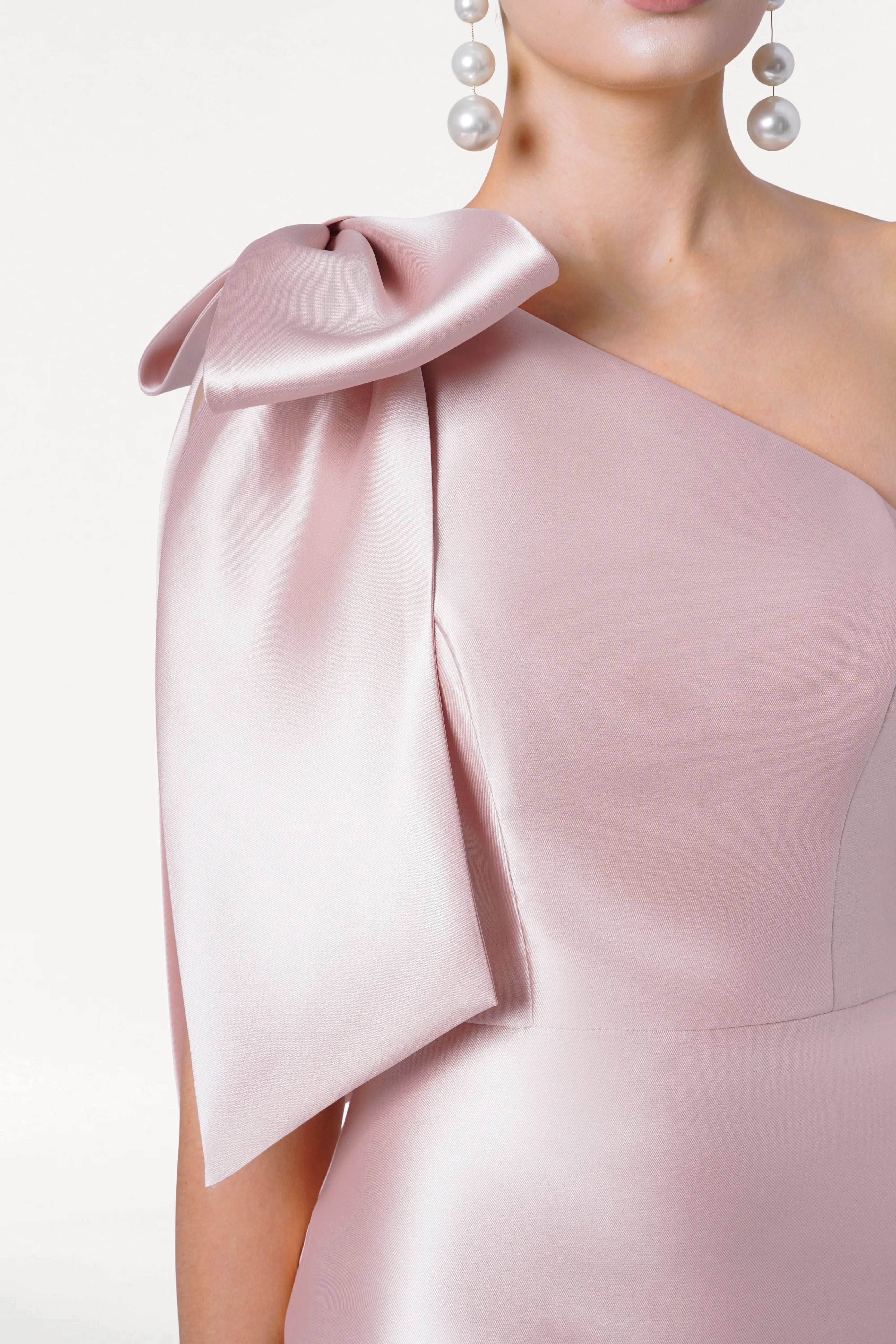 A woman models the TH&TH Ritz Bridesmaid Dress in blush pink satin twill, featuring a one-shoulder design with a statement bow. She wears dangling pearl earrings. The image is cropped at chest level, omitting her full face.