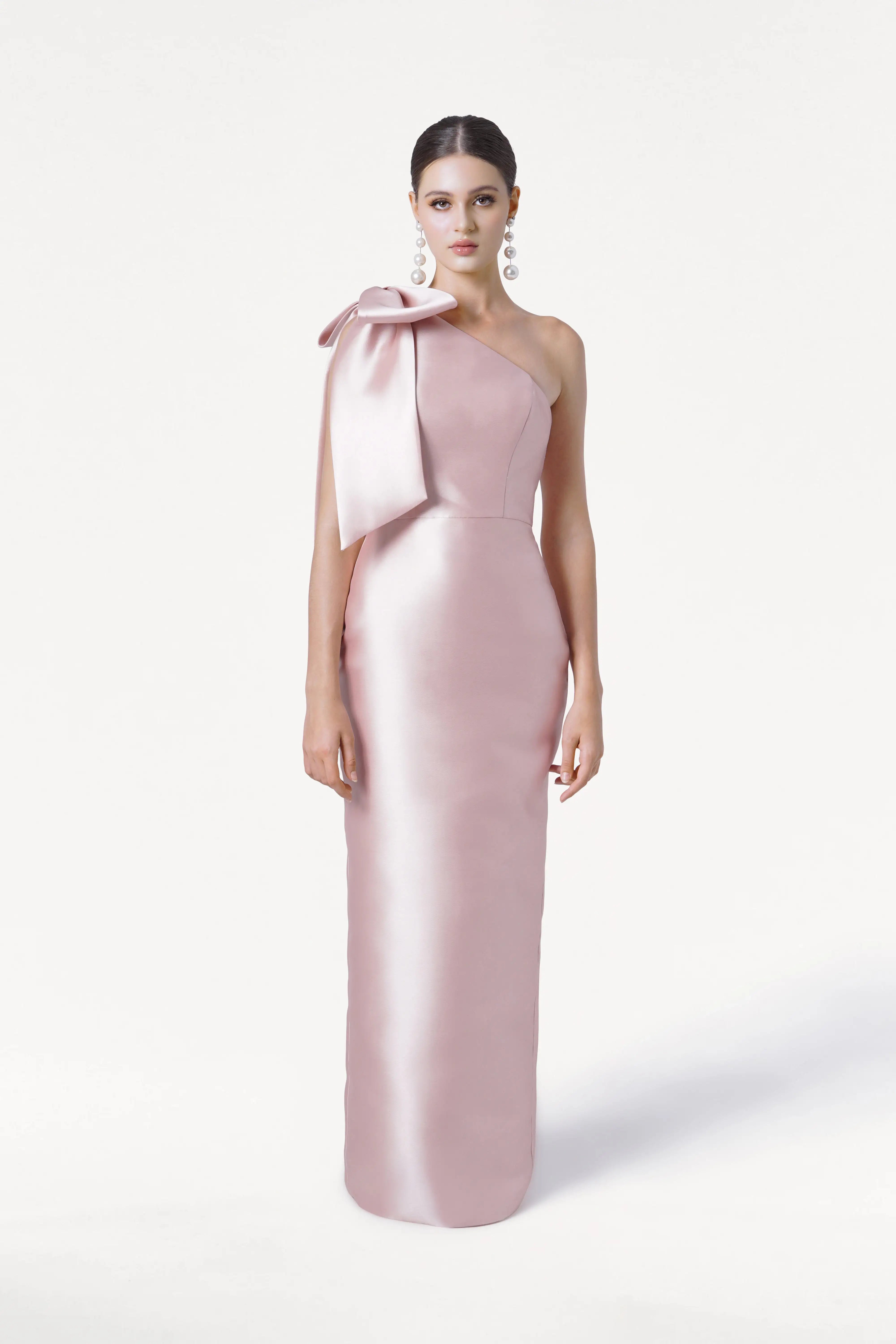 A woman wears the TH&TH Ritz Bridesmaid Dress in blush pink satin twill, featuring a one-shoulder design with a statement bow. She stands against a white background, her hair in an updo and styled with elegant dangling earrings.