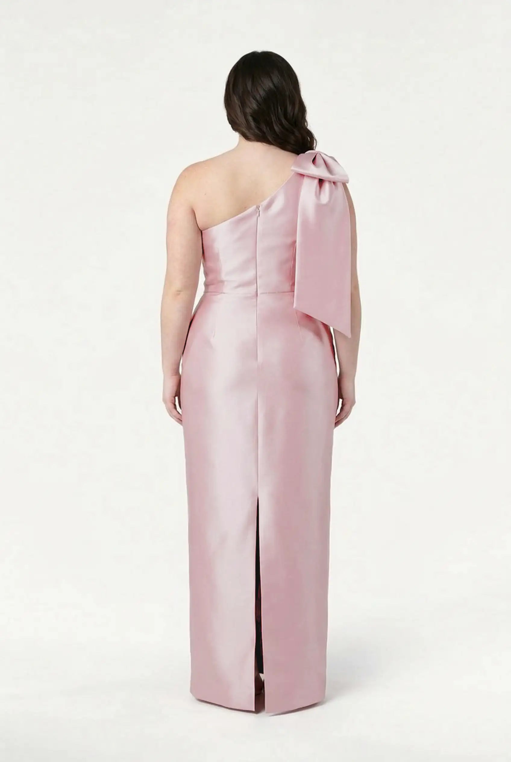 A woman wearing the TH&TH Ritz Bridesmaid Dress in Blush Pink Satin Twill stands facing away, highlighting the elegant back and statement shoulder bow, set against a plain white background.
