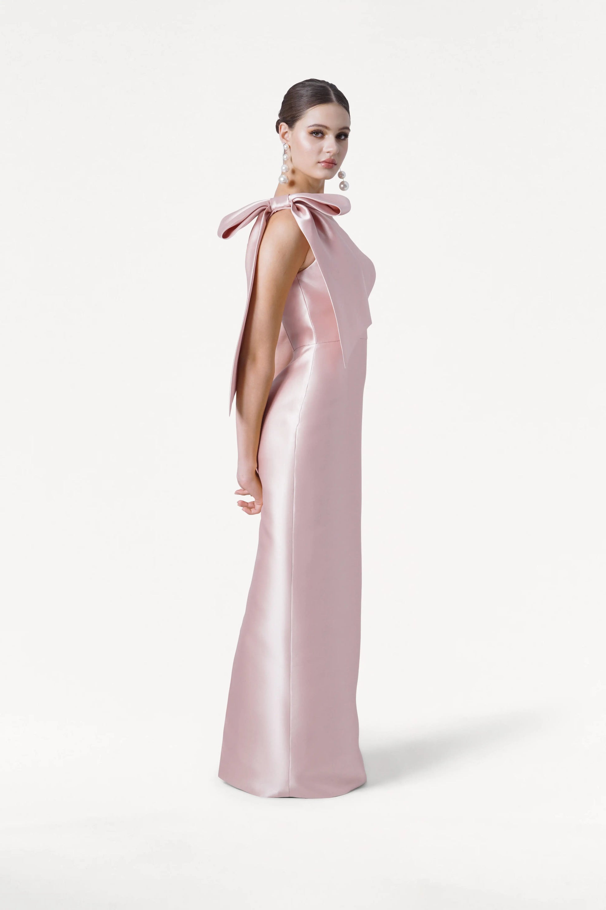 A woman in profile wears the TH&TH Ritz Bridesmaid Dress in Blush Pink Satin Twill, a long one-shoulder gown with a statement bow, paired with pearl earrings and a sleek bun against a plain white background.