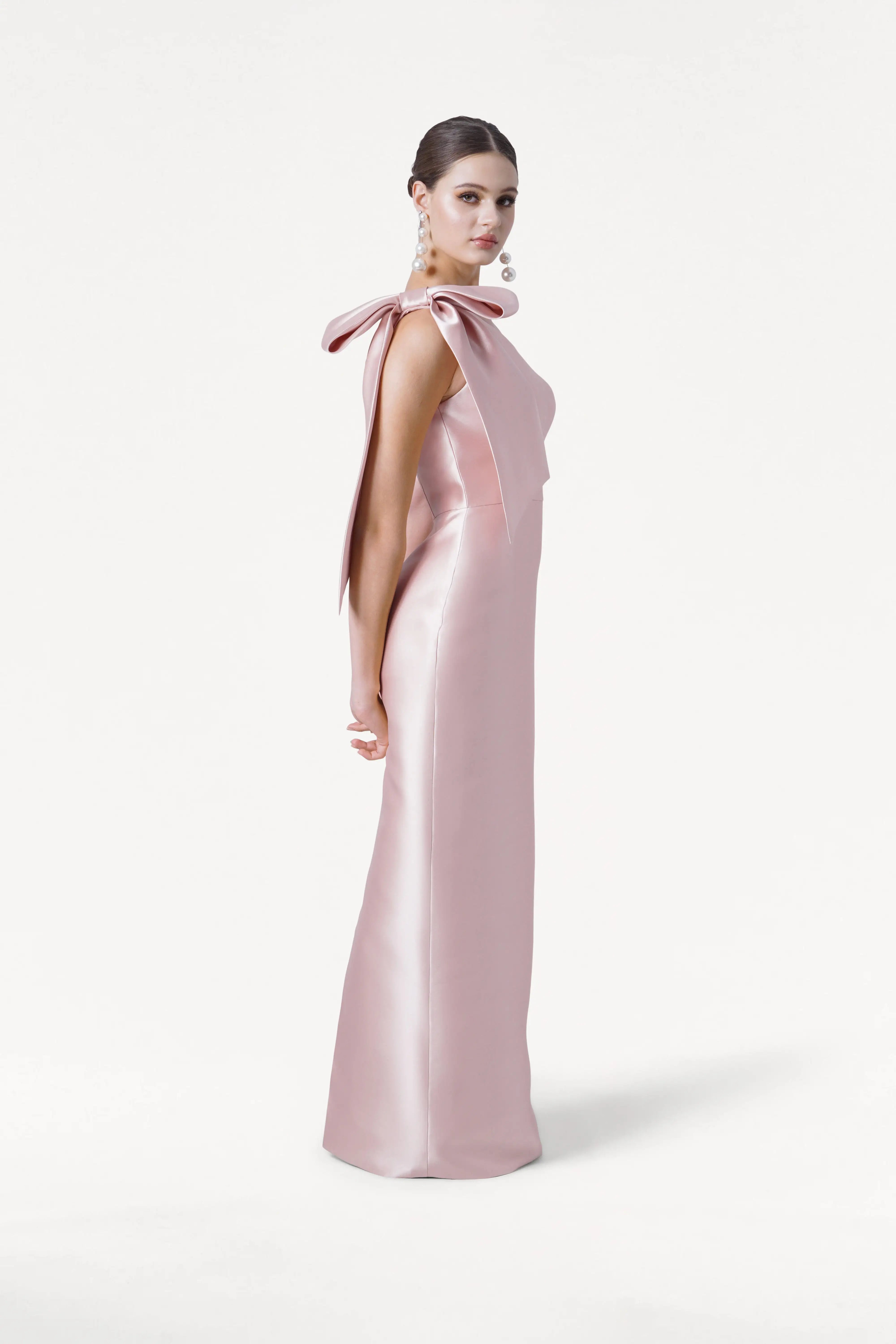 A woman in profile wears the TH&TH Ritz Bridesmaid Dress in Blush Pink Satin Twill, a long one-shoulder gown with a statement bow, paired with pearl earrings and a sleek bun against a plain white background.