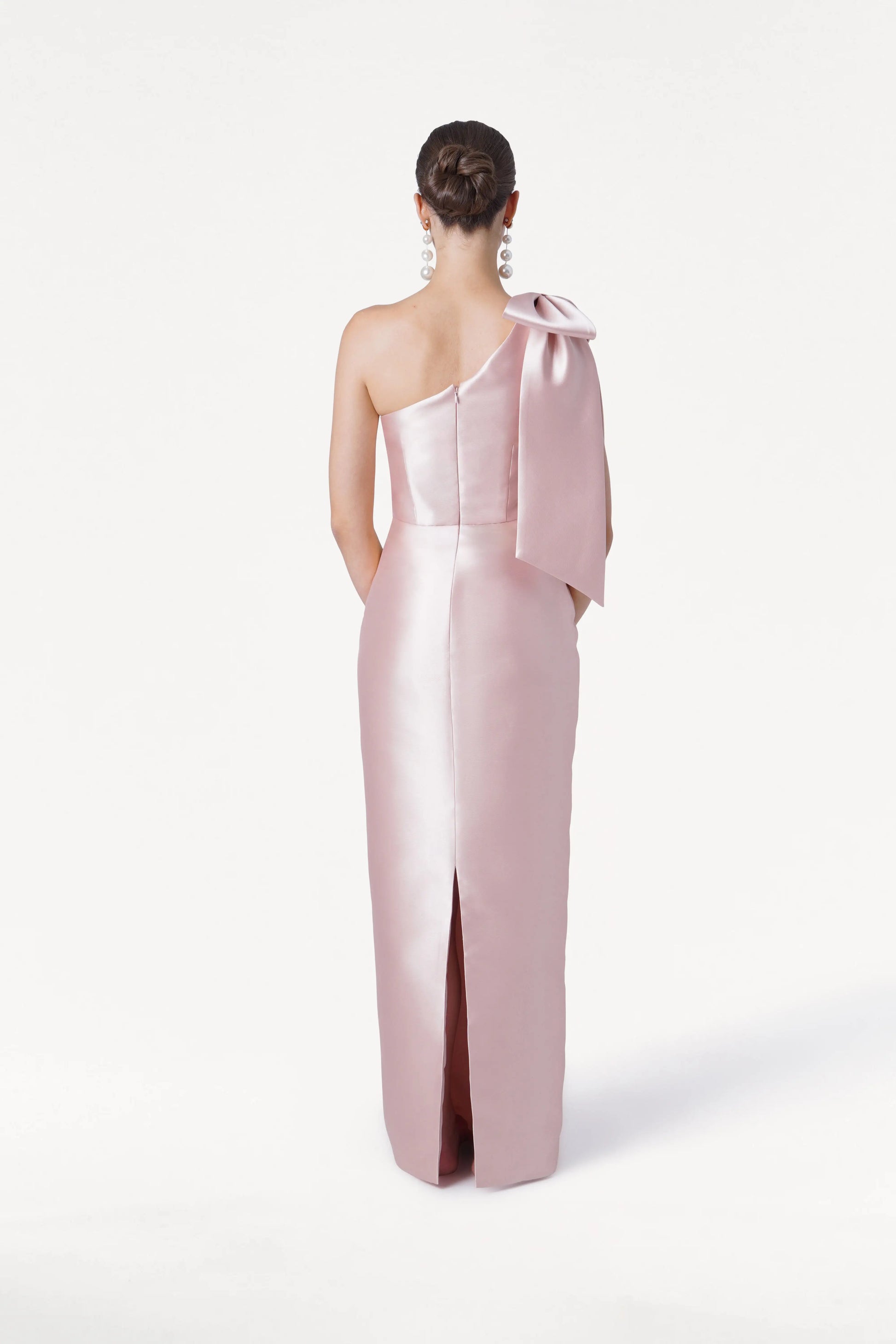 A woman with her hair in a low bun and large dangling earrings faces away, wearing the TH&TH Ritz Bridesmaid Dress in blush pink satin twill—a one-shoulder style with a statement bow and back slit. The background is plain white.