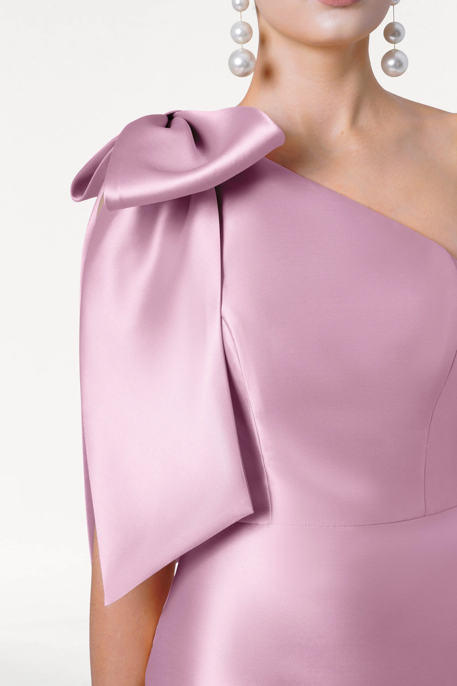 A woman wears the TH&TH Ritz Bridesmaid Dress in Bubblegum Pink Satin Twill, featuring a statement one-shoulder bow and pearl drop earrings, shown from the chest up against a white background.