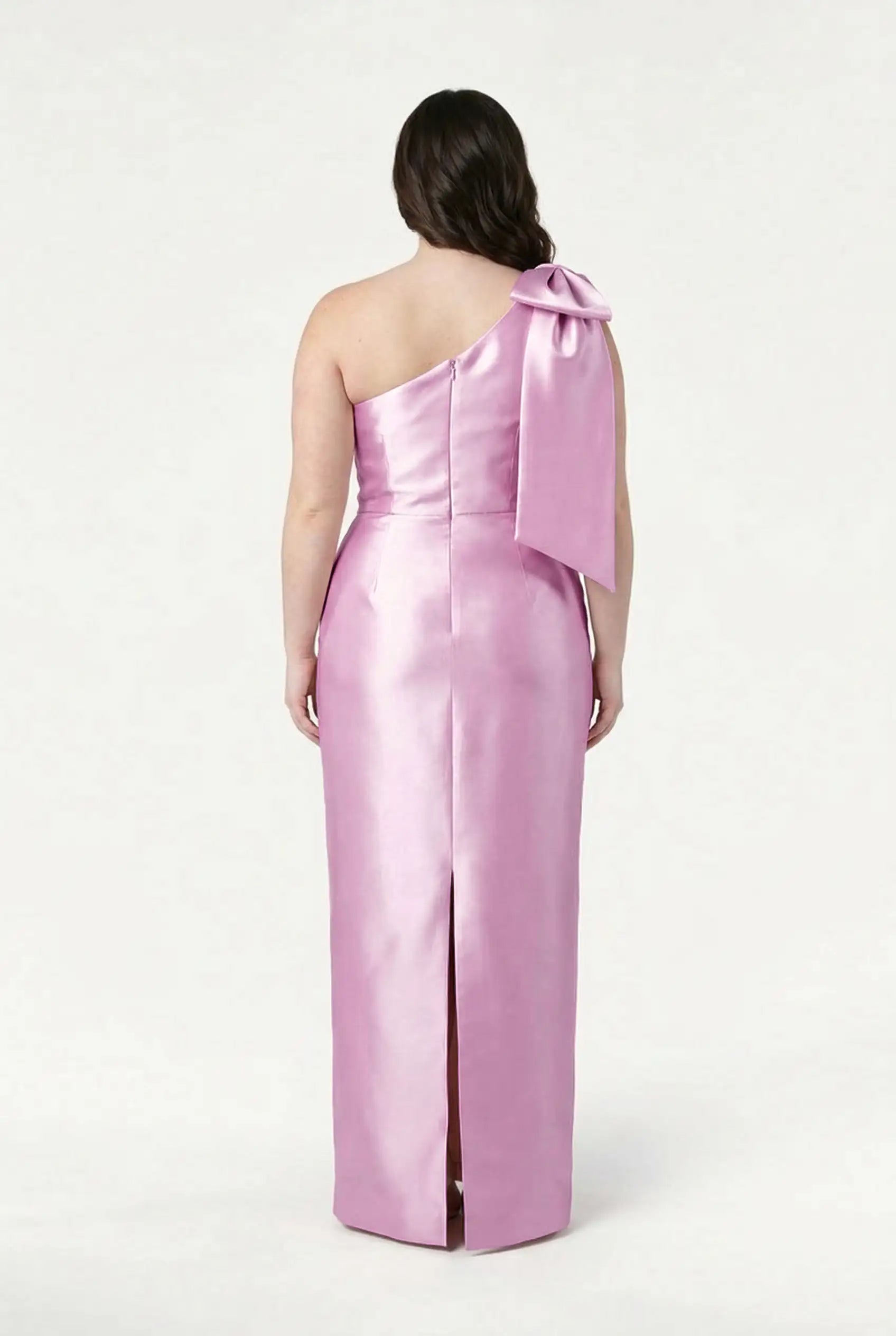 A woman faces away in the TH&TH Ritz Bridesmaid Dress in Bubblegum Pink Satin Twill, featuring a shiny one-shoulder design with a statement bow and center back slit. The plain white background highlights the gown.