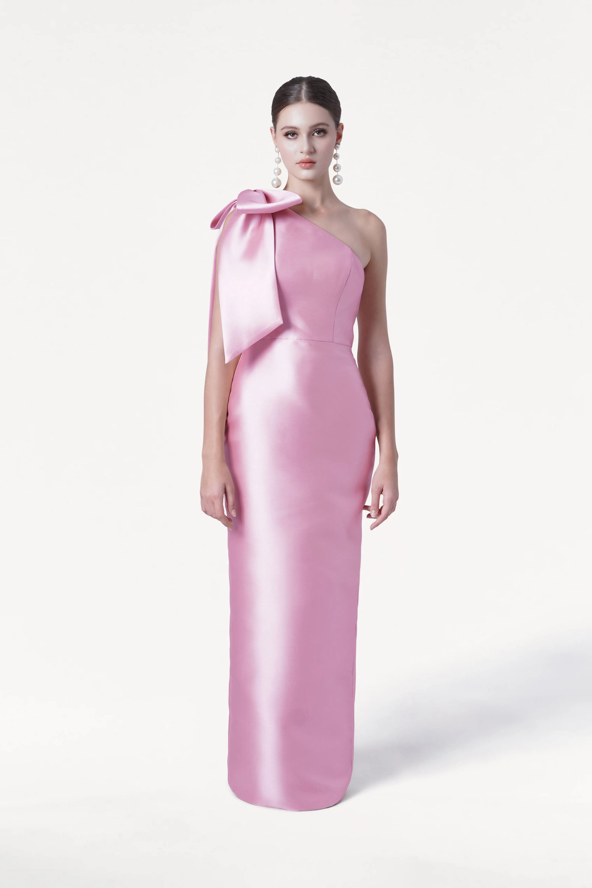 A woman stands against a plain white background wearing the TH&TH Ritz Bridesmaid Dress in Bubblegum Pink Satin Twill, a shiny, floor-length one-shoulder gown with a statement bow on the shoulder. She pairs it with statement drop earrings and has her hair pulled back.