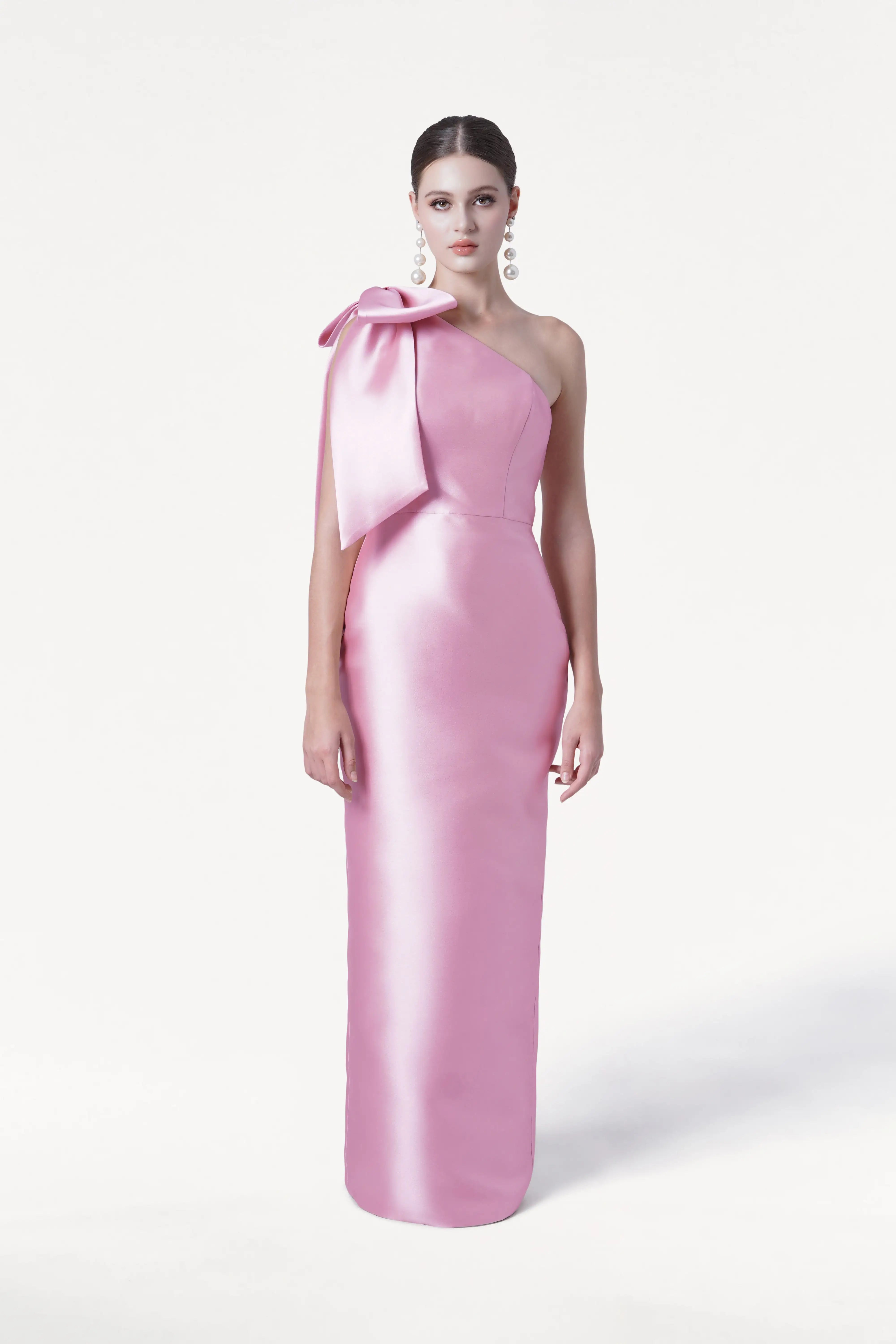 A woman stands against a plain white background wearing the TH&TH Ritz Bridesmaid Dress in Bubblegum Pink Satin Twill, a shiny, floor-length one-shoulder gown with a statement bow on the shoulder. She pairs it with statement drop earrings and has her hair pulled back.