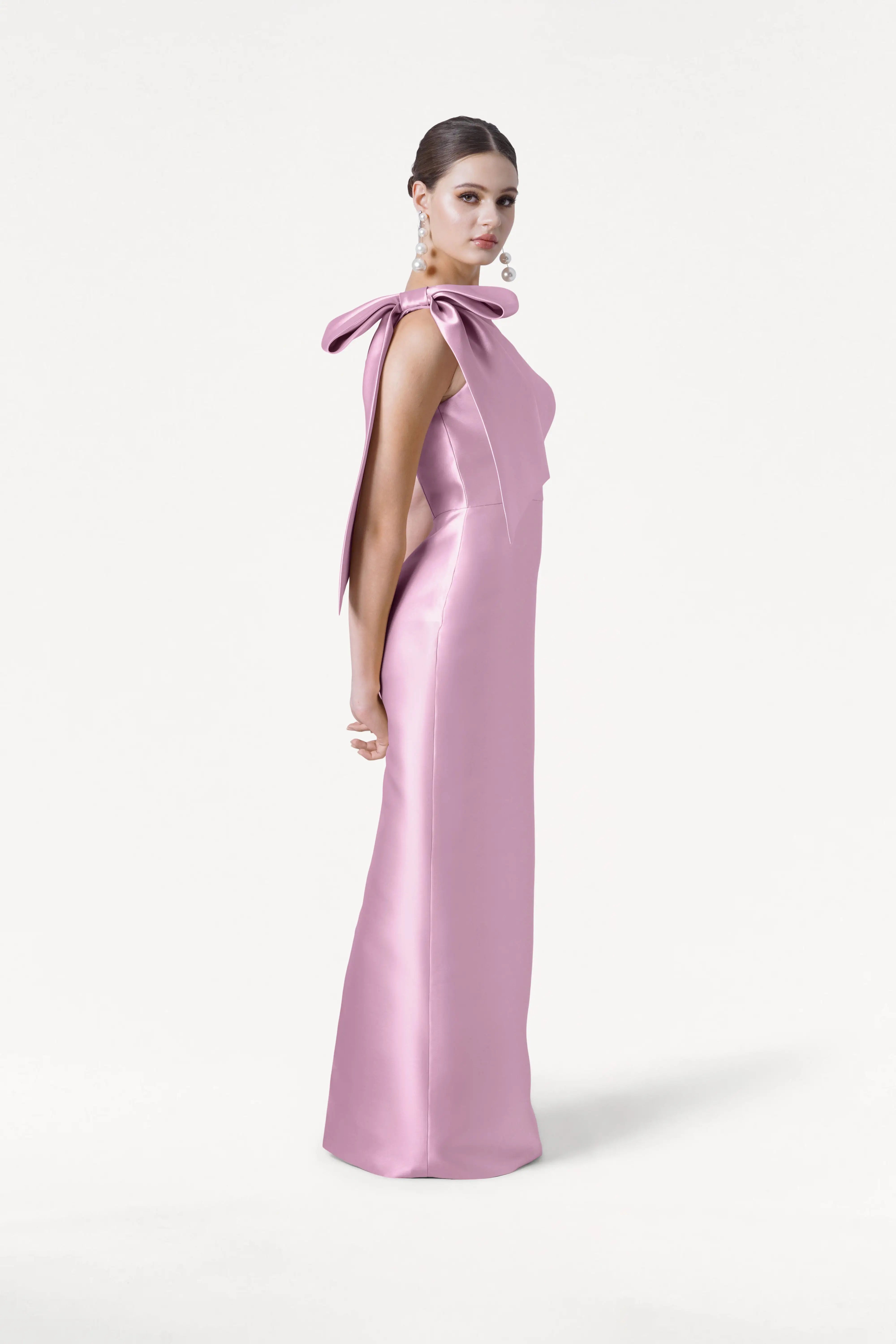 A woman stands against a white background wearing the TH&TH Ritz Bridesmaid Dress in Bubblegum Pink Satin Twill, featuring a floor-length silhouette and statement one-shoulder bow, paired with statement earrings and a neat bun.
