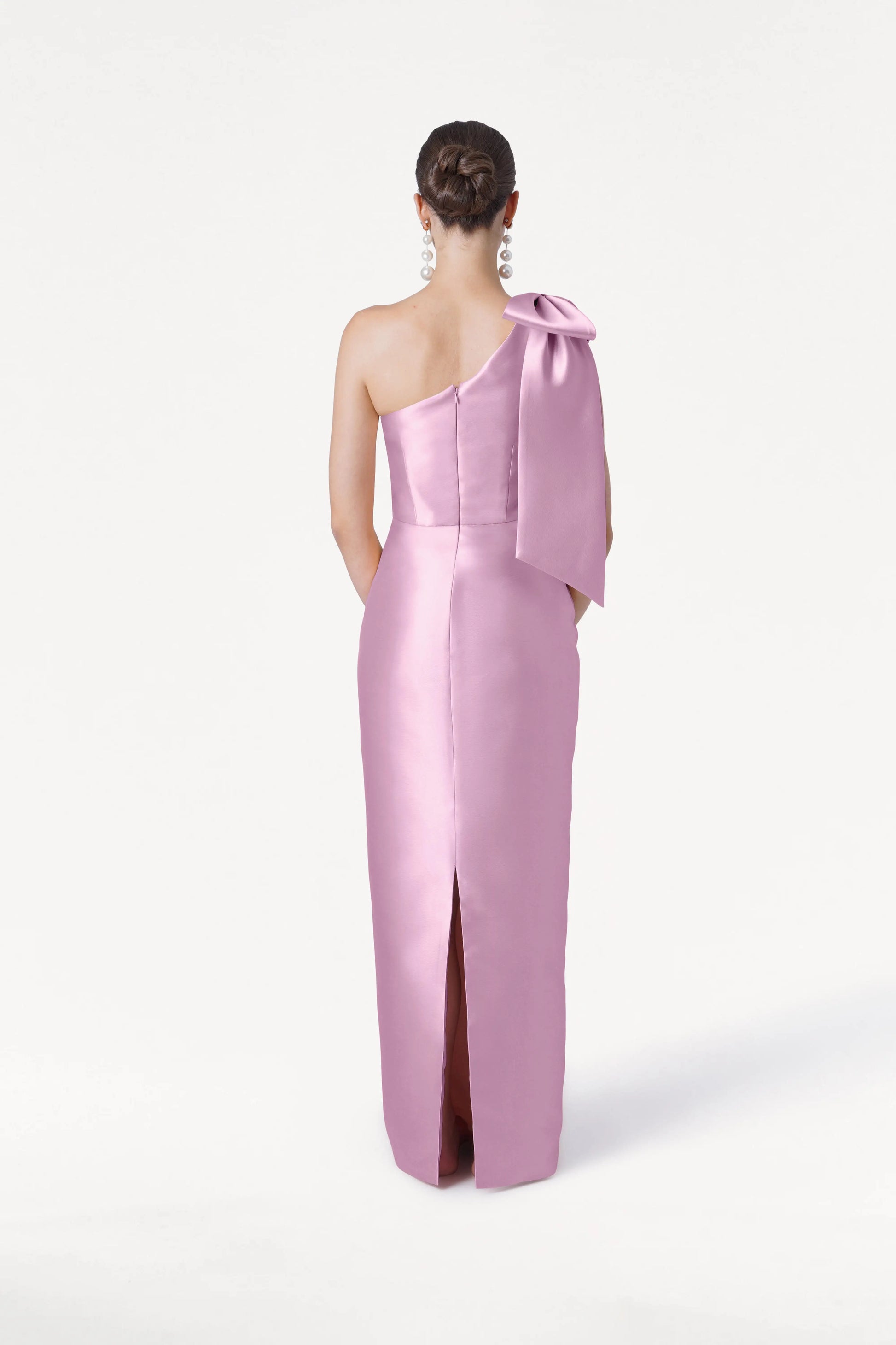 A woman with her hair in a neat bun and dangling earrings faces away, wearing the TH&TH Ritz Bridesmaid Dress in Bubblegum Pink Satin Twill, featuring a one-shoulder design with statement bow and back slit, set against a plain white background.