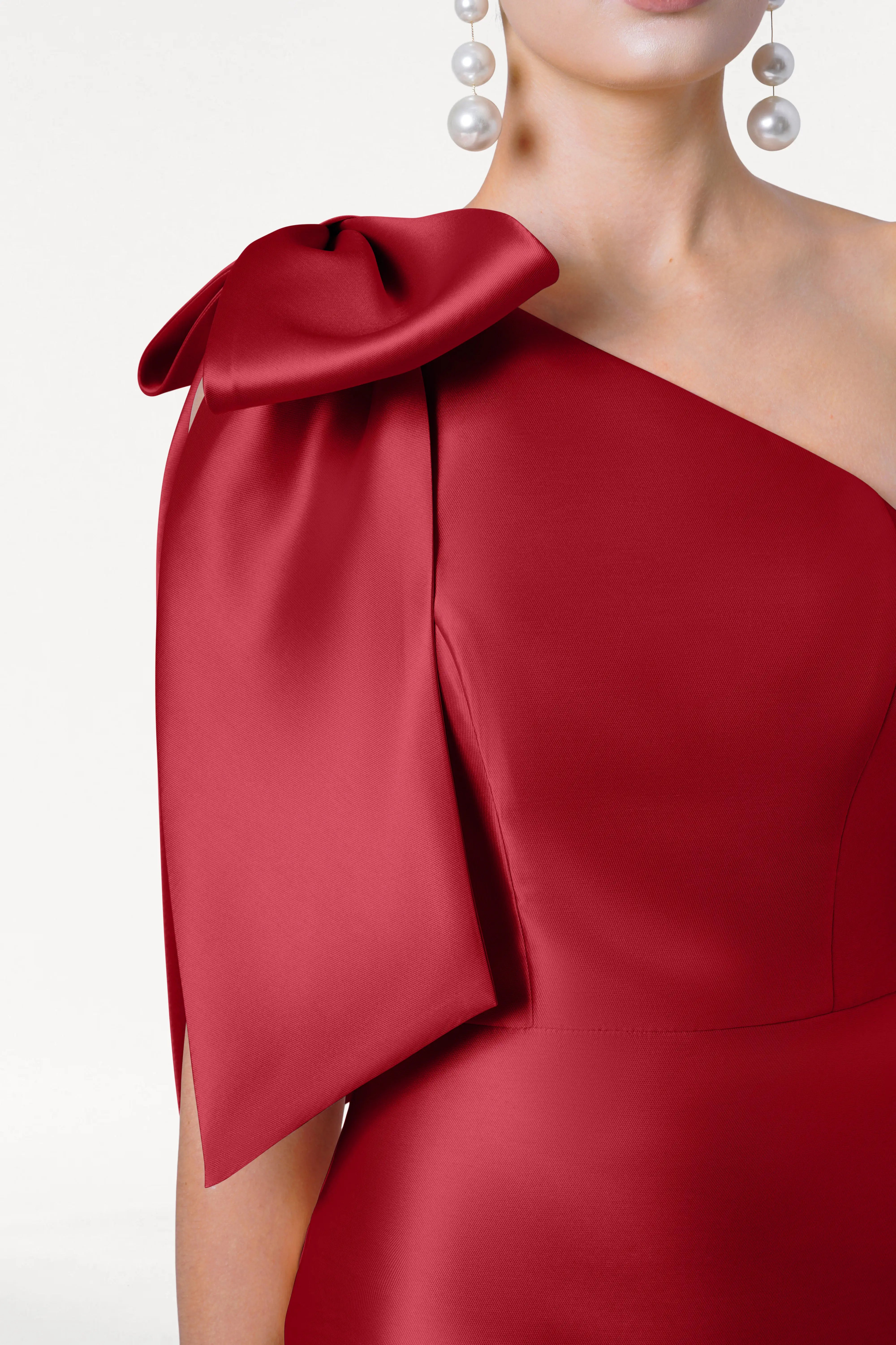 A woman wears the TH&TH Ritz Bridesmaid Dress in Dark Cherry Satin Twill, featuring a one-shoulder design with a large bow. She pairs it with dangling pearl earrings and stands against a white background.